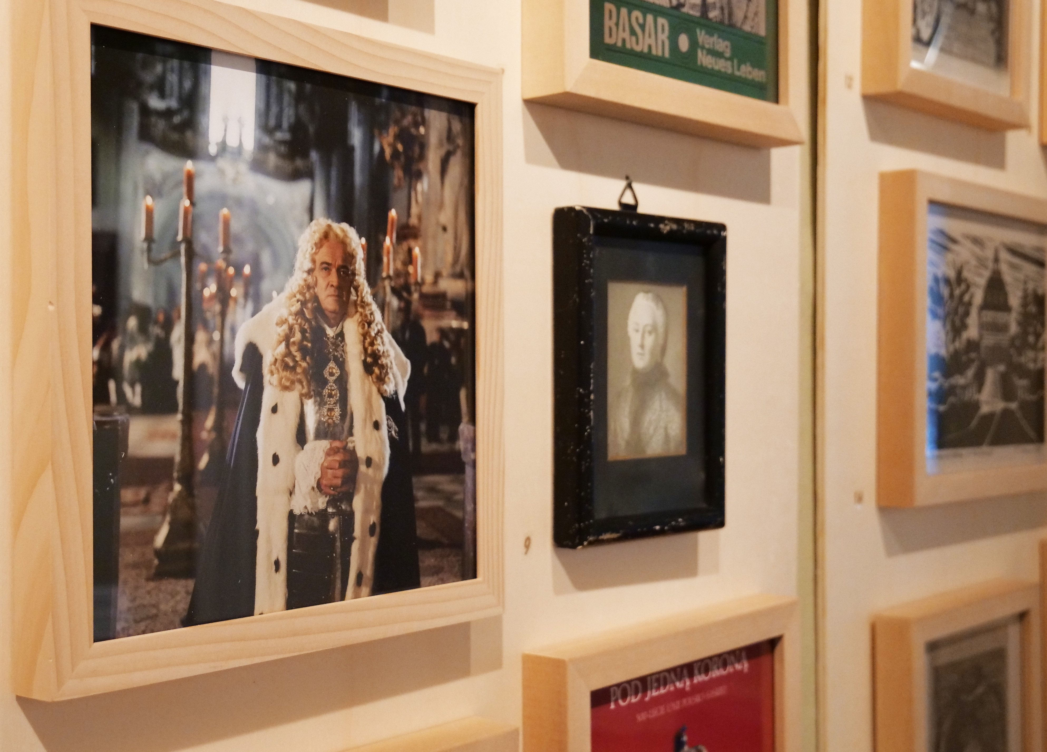 KI generiert: Das Bild zeigt eine Wand mit einer Sammlung von gerahmten Bildern und Plakaten, die wahrscheinlich historische oder künstlerische Themen darstellen. Links ist ein gelbes Plakat mit der Aufschrift "Mit Glanz & Gloria" zu sehen.