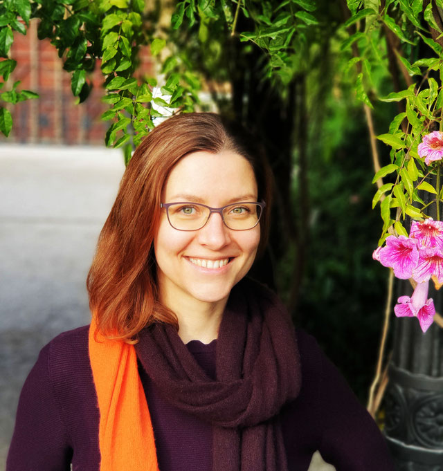 KI generiert: Das Bild zeigt eine lächelnde Person mit Brille, die von grünen Blättern und rosa Blüten umgeben ist. Die Person trägt einen schwarzen Schal und einen orangefarbenen Akzent, was einen Kontrast zur natürlichen Umgebung bildet.