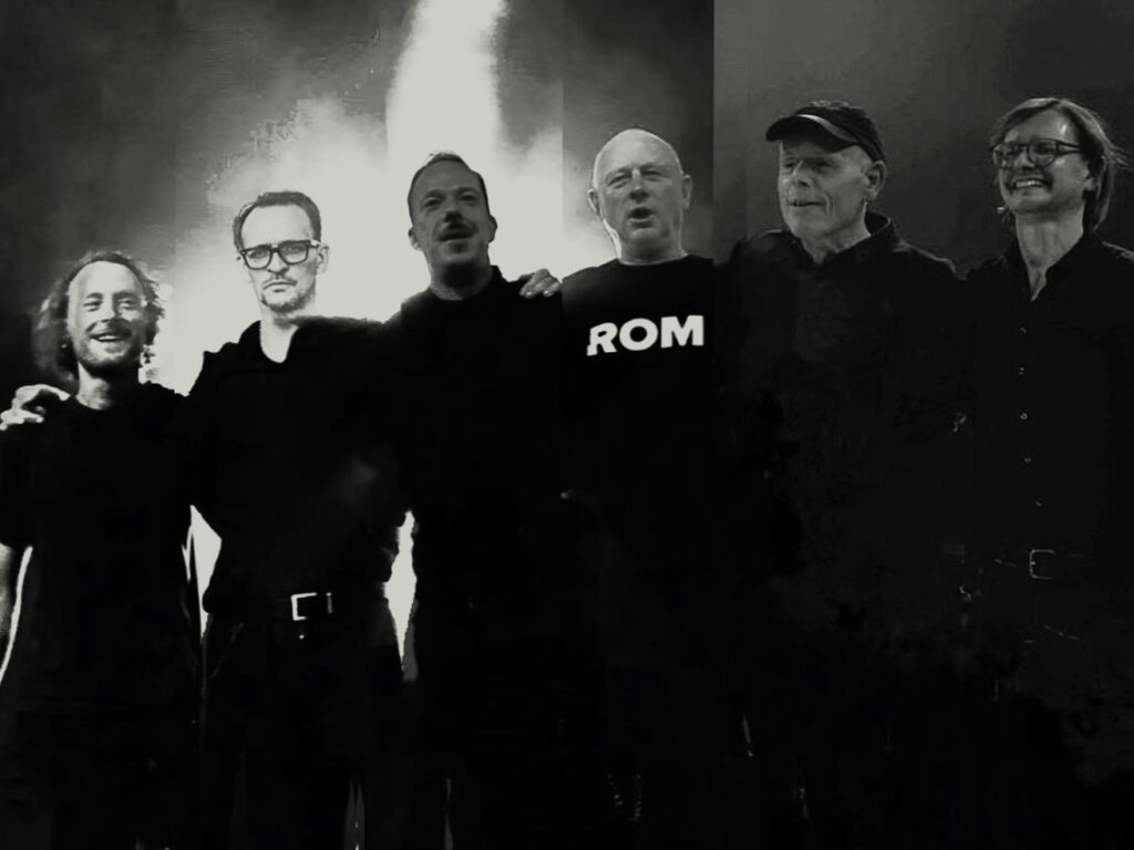 KI generiert: Das Bild zeigt sechs Personen in schwarzer Kleidung, die nebeneinander vor einem Bühnenhintergrund stehen. Sie wirken harmonisch und scheinen gemeinsam auf einer Veranstaltung oder bei einem Auftritt zu sein.