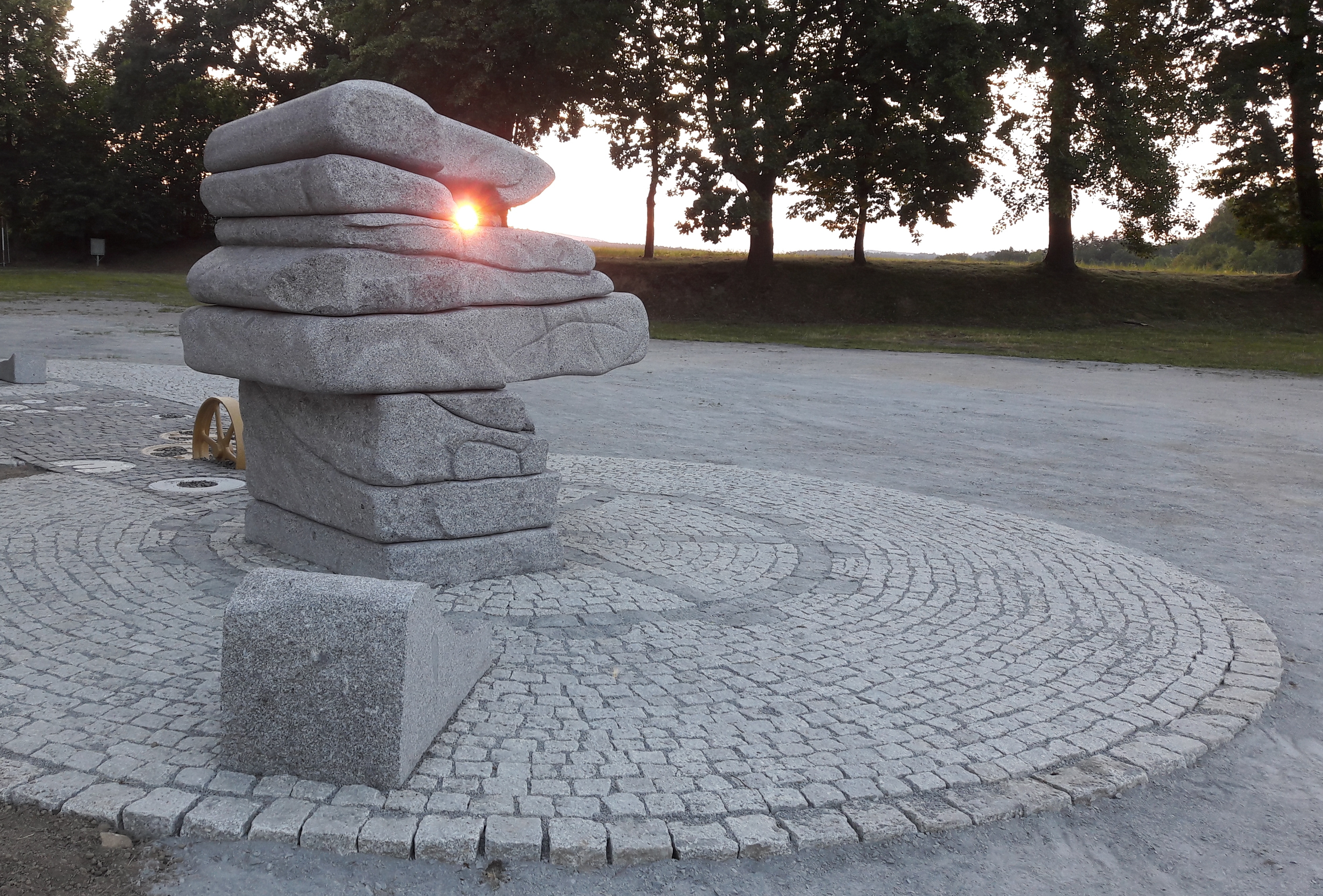 KI generiert: Das Bild zeigt eine Steinskulptur, bei der die Sonne durch einen Spalt zwischen den Steinen scheint. Die Skulptur ist von einem gepflasterten Kreis umgeben, und im Hintergrund sind Bäume zu sehen.