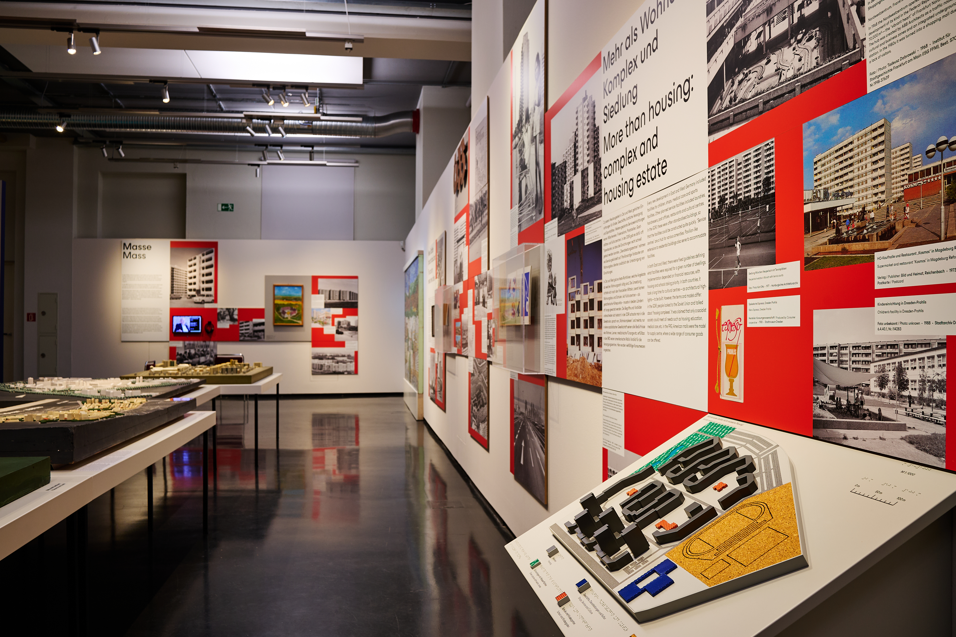 KI generiert: Das Bild zeigt eine Ausstellung in einem Museum, die sich mit Architektur und Wohnkomplexen befasst. Es sind verschiedene Modelle, Fotos und Informationstafeln zu sehen, die das Thema illustrieren.