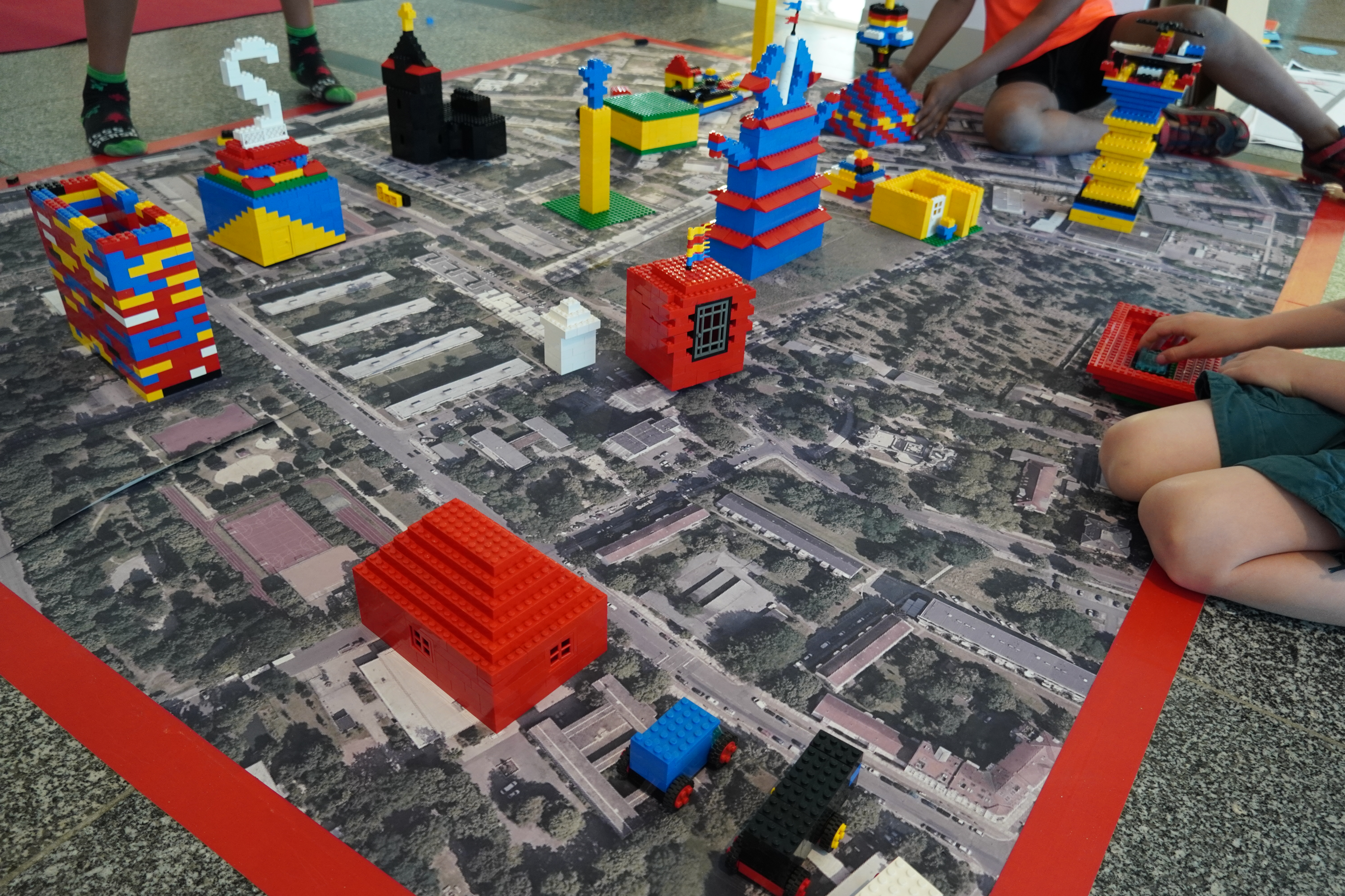 KI generiert: Das Bild zeigt Kinder, die verschiedene bunte Gebäude aus Legosteinen auf einer Karte einer Stadt oder eines Stadtteils bauen. Der Hauptinhalt ist die kreative Gestaltung einer urbanen Landschaft mit Legosteinen.