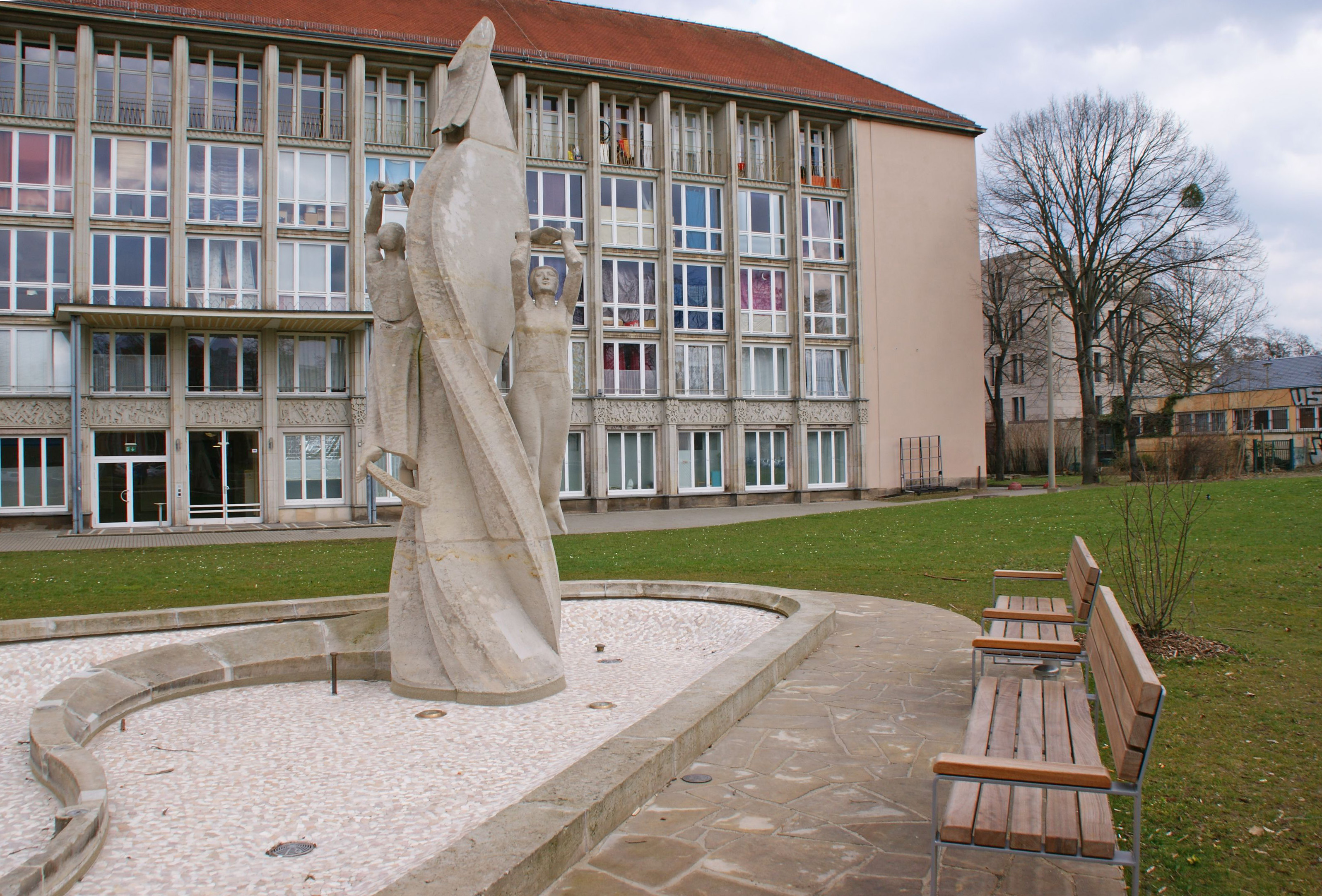 AI generated: The image shows a modern stone sculpture group placed in front of a building with several windows and a green area. Next to it are some benches on a paved path.