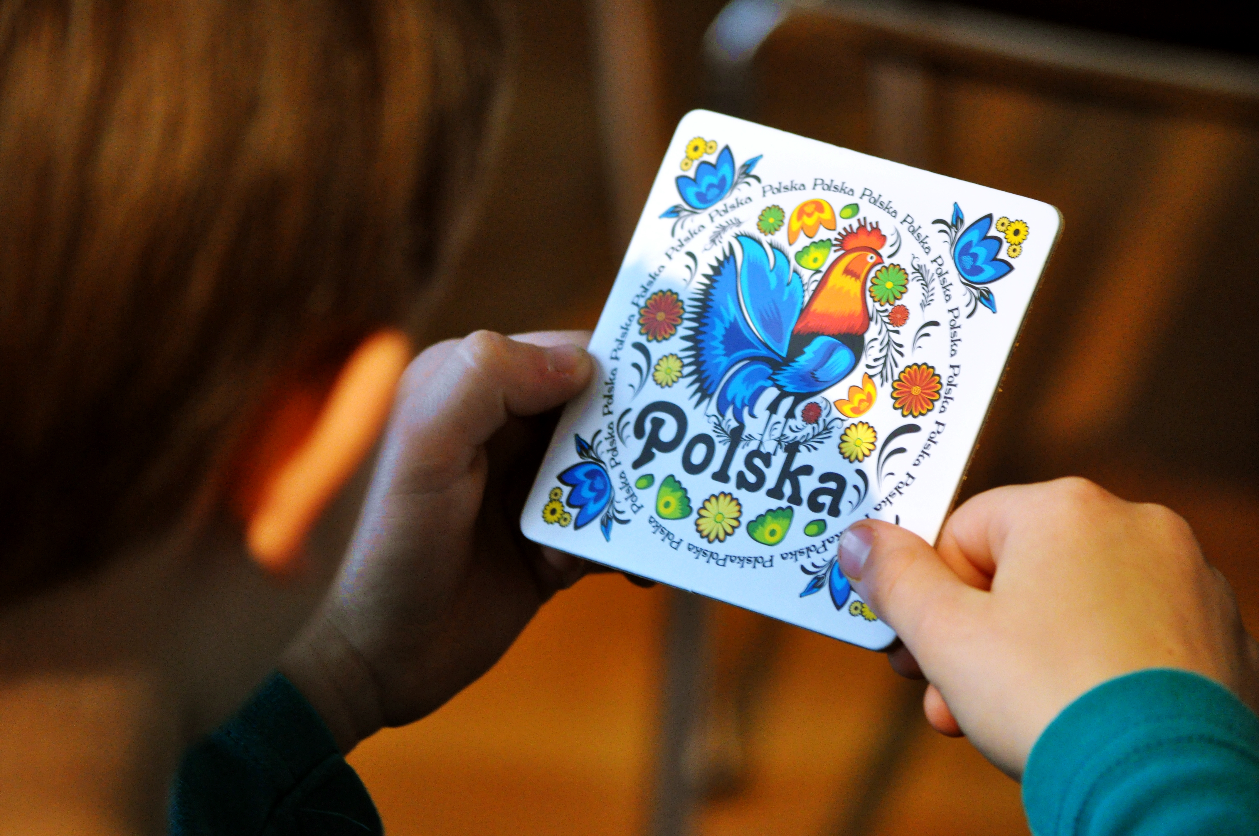 KI generiert: Ein Kind hält eine Karte mit einer bunten Illustration eines Hahns, Blumen und der Aufschrift "Polska" in den Händen. Die Karte weist traditionelle polnische Motive auf.