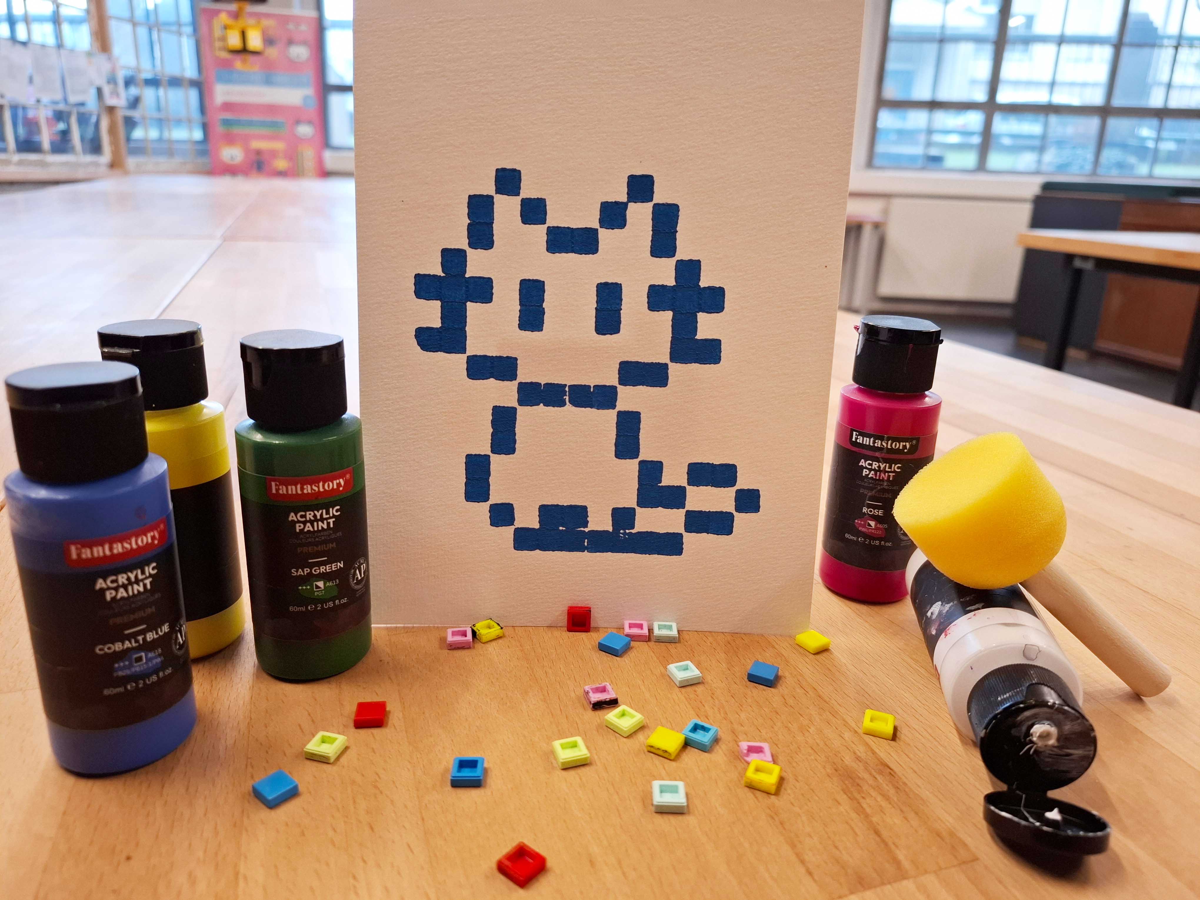 KI generiert: Auf dem Bild ist ein Leinwandgemälde mit einer pixelartigen, blauen Katzenfigur zu sehen, umgeben von Acrylfarben und bunten Mosaiksteinen auf einem Holztisch. Der Hauptinhalt des Bildes ist die kreative Darstellung dieser digitalen Katze.