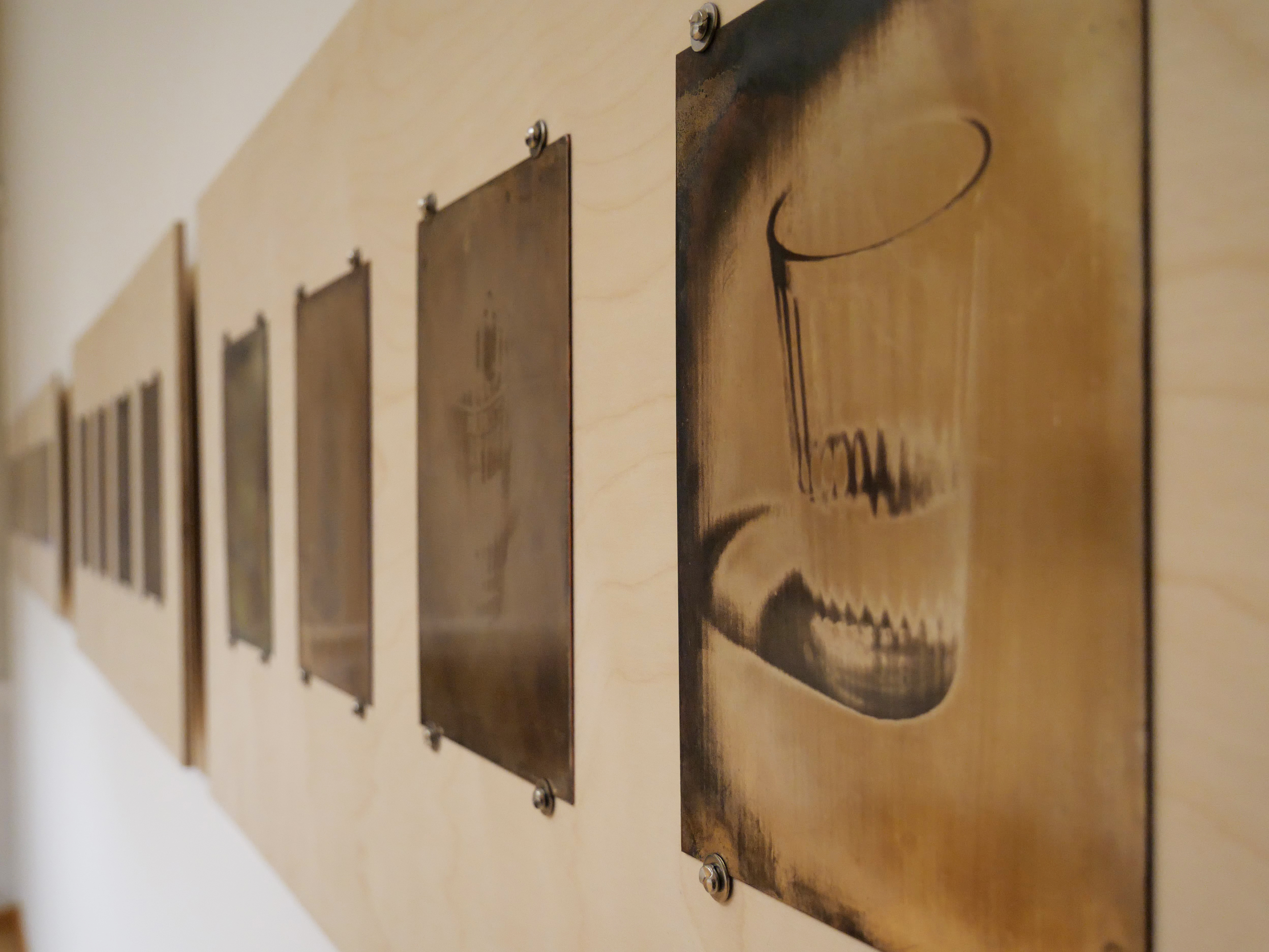 KI generiert: Das Bild zeigt eine Anordnung von metallischen Platten mit eingravierten Motiven, die an einer Holzwand befestigt sind. Im Vordergrund ist ein Gravurbild eines Glases zu erkennen.