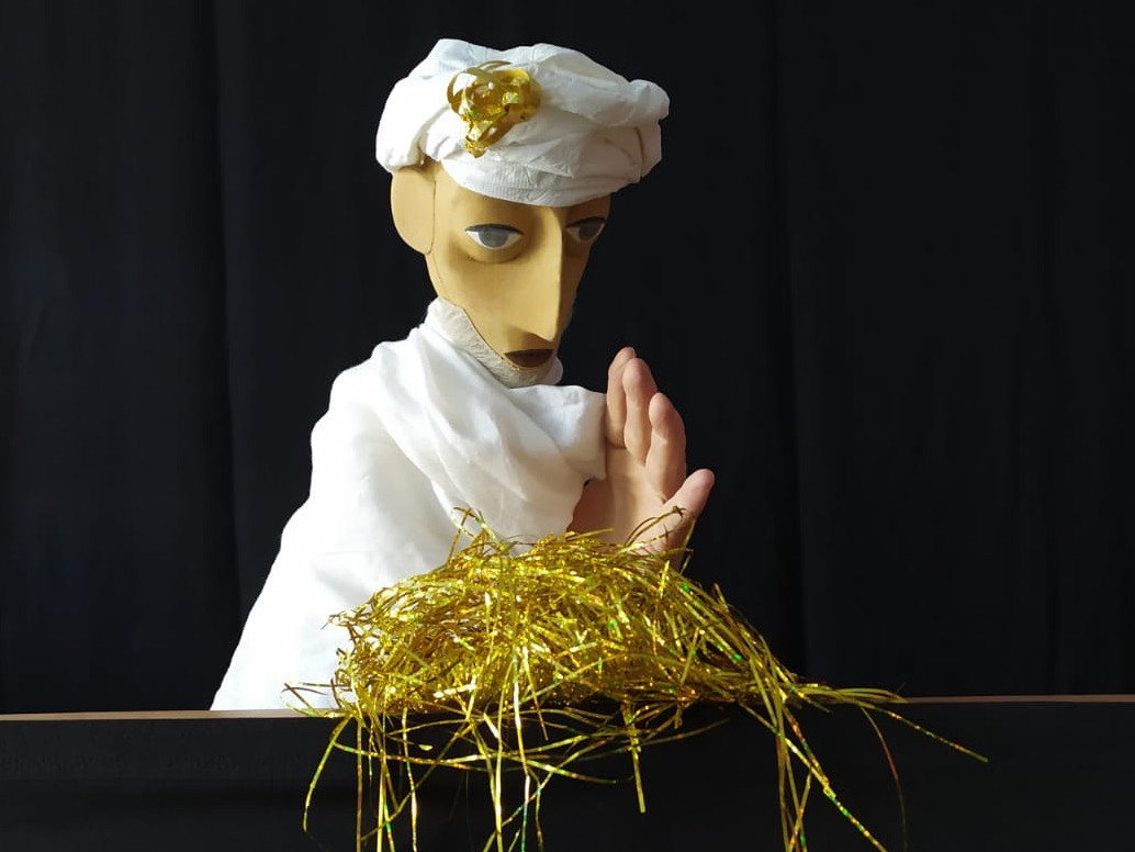 KI generiert: Das Bild zeigt eine Puppe mit einem weißen Turban und Gewand, die hinter einem Tisch mit goldenen Fäden steht. Der Hintergrund ist schwarz, was die Puppe in den Mittelpunkt rückt.