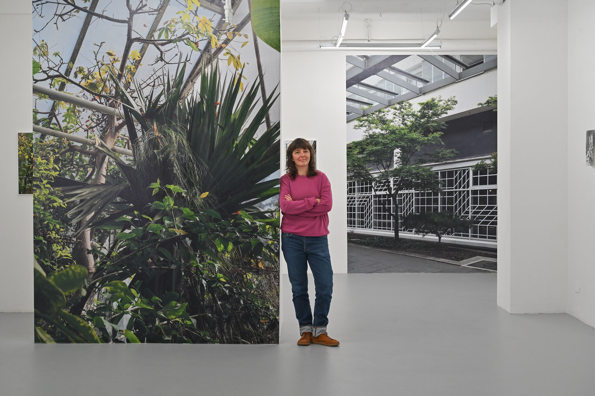 KI generiert: Das Bild zeigt eine Person in einem Galerie- oder Ausstellungsraum, die vor großen Wandinstallationen mit Naturmotiven und einem modernen Gebäude posiert. Der Raum wirkt modern und minimalistisch, mit hellen Wänden und moderner Beleuchtung.