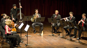 KI generiert: Das Bild zeigt ein Kammermusikensemble, das auf einer Bühne spielt. Die Musiker sind mit verschiedenen Instrumenten wie Cello, Kontrabass, Horn, Flöte und Oboe ausgestattet.