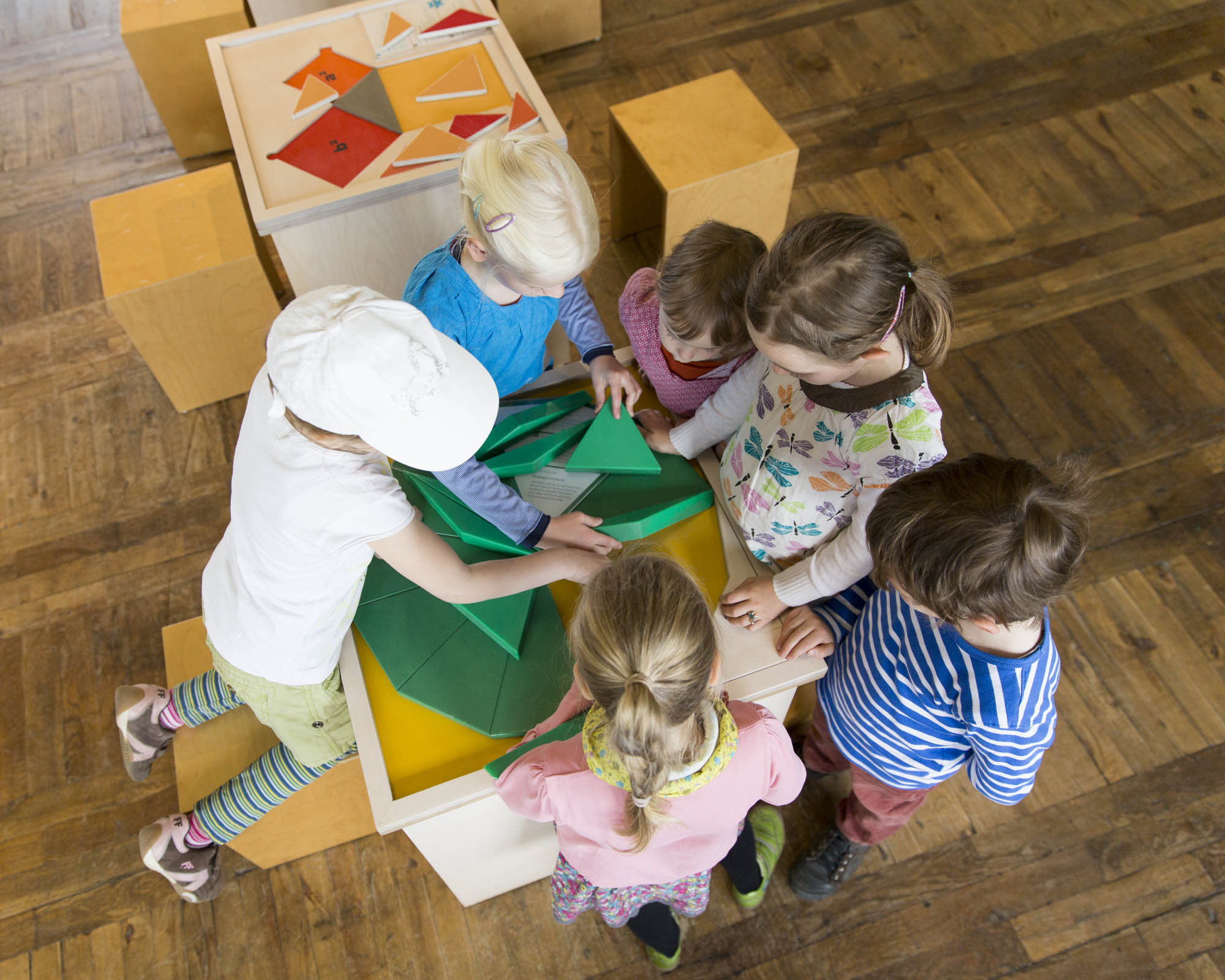 KI generiert: Das Bild zeigt eine Gruppe von Kindern, die um einen Tisch herum stehen und gemeinsam mit bunten geometrischen Formen spielen. Sie scheinen in eine kreative oder lernorientierte Aktivität vertieft zu sein.