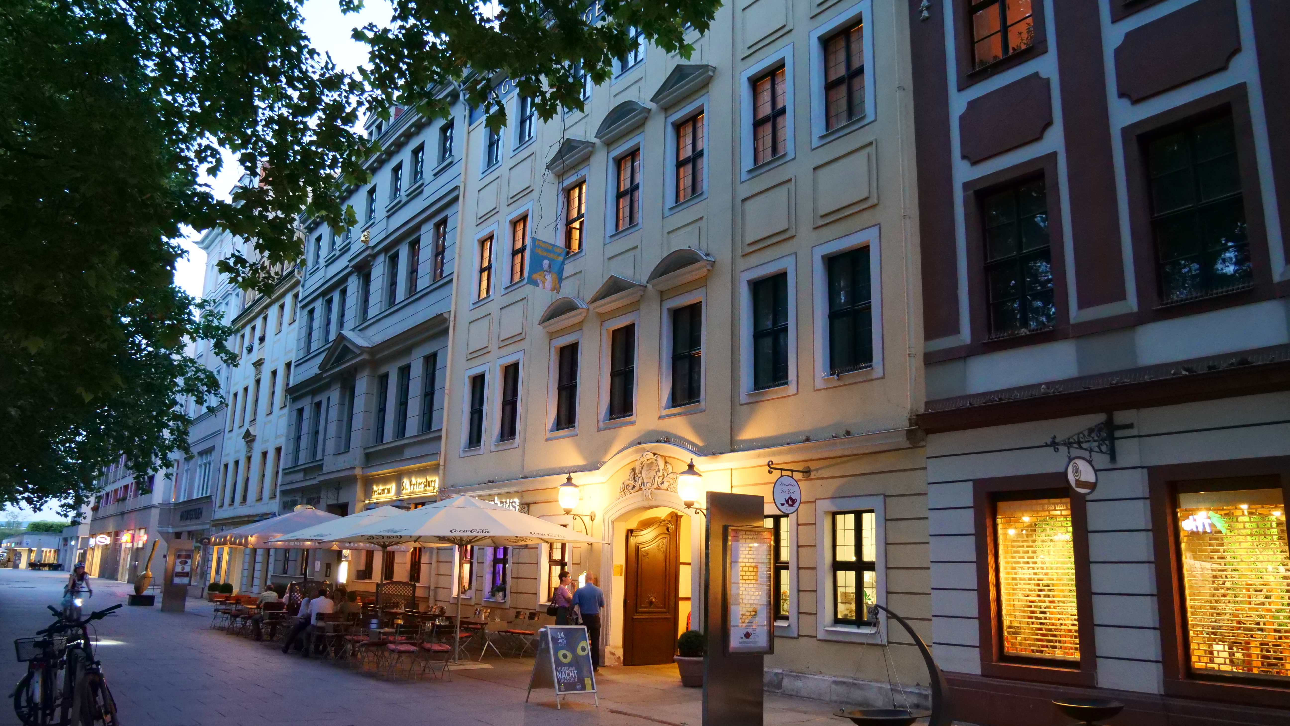 KI generiert: Das Bild zeigt eine Reihe historischer Gebäude mit einem Restaurant oder Café im Erdgeschoss, dessen Außenbereich mit Tischen und Sonnenschirmen ausgestattet ist. Die Straße ist ruhig und menschenleer, bis auf einige vereinzelte Passanten.