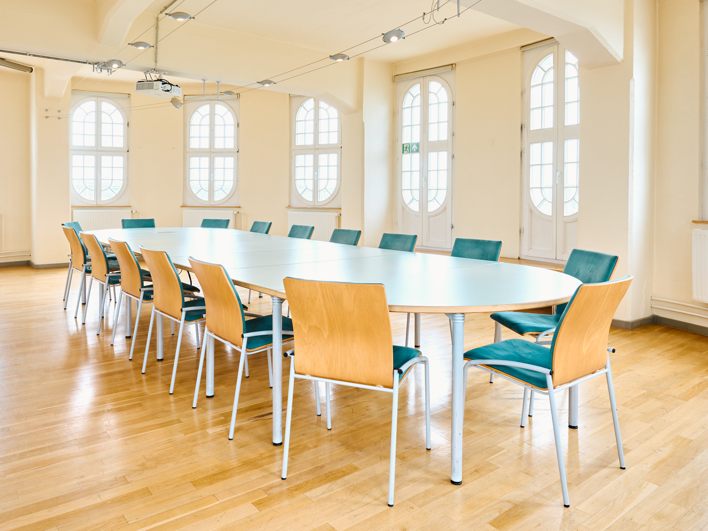 KI generiert: Das Bild zeigt einen Konferenzraum mit einem ovalen Tisch, um den 12 Stühle angeordnet sind. Der Raum ist hell und freundlich gestaltet, mit großen Fenstern und Holzboden.