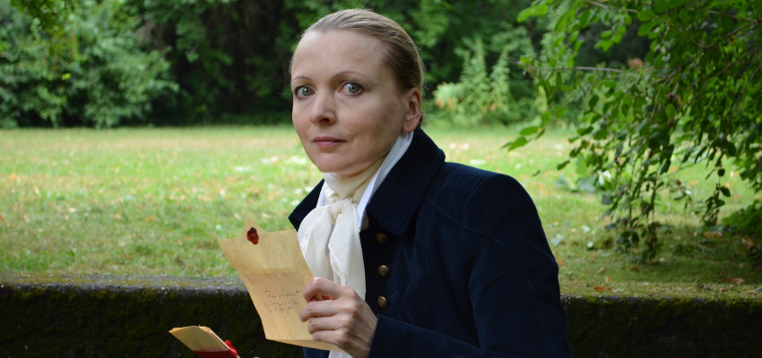 KI generiert: Das Bild zeigt eine Person in historischer Kleidung, die draußen ist und ein Schriftstück in der Hand hält. Im Hintergrund ist eine grüne Landschaft erkennbar.