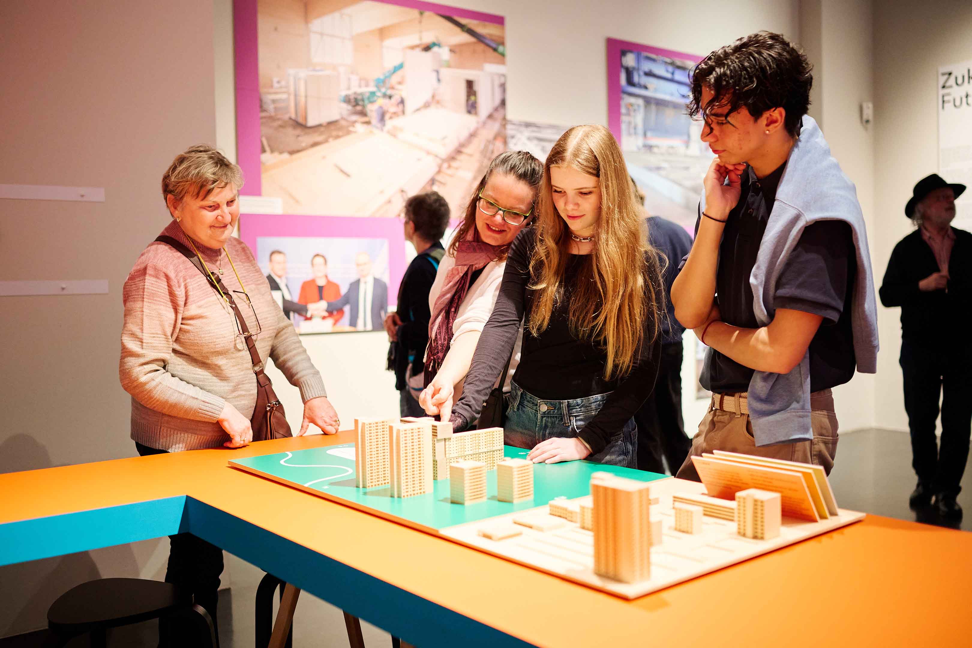 AI generated: The image shows a group of people looking at an architectural model with high-rise buildings. In the background you can see pictures on the wall that match the theme of the exhibition.