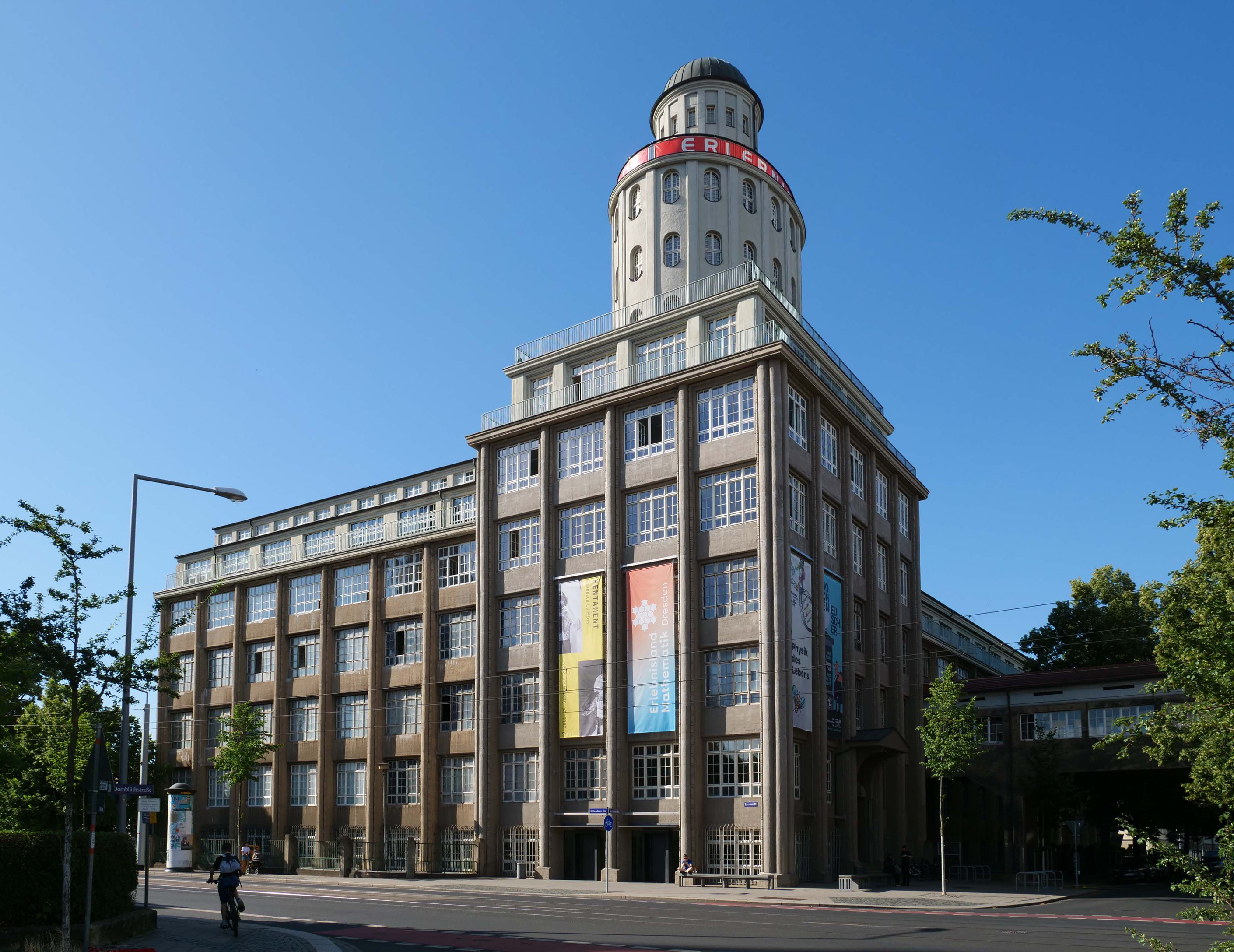 KI generiert: Das Bild zeigt ein großes, mehrstöckiges Gebäude mit einem markanten runden Turm an der Spitze, auf dem "FRIGOR" steht. Es ist umgeben von Bäumen und beleuchtet von Sonnenlicht, mit einem klaren blauen Himmel im Hintergrund.