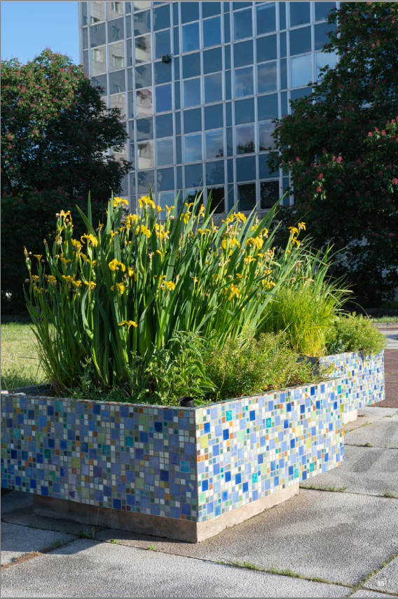 KI generiert: Das Bild zeigt ein farbenfrohes Mosaik-Beet, das mit gelben Blumen und grünem Laub bepflanzt ist. Im Hintergrund ist ein modernes Glasgebäude zu sehen.