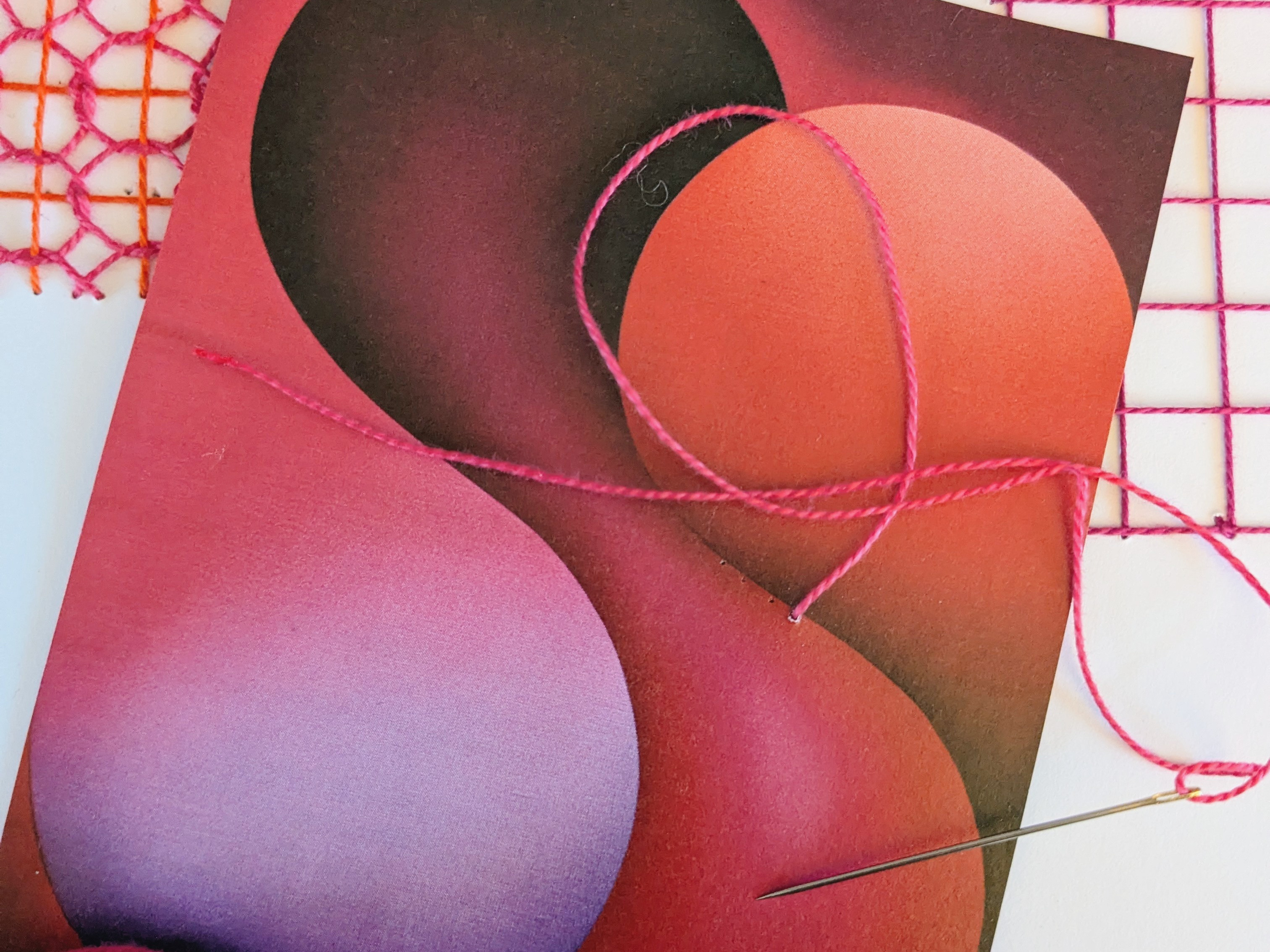 KI generiert: Das Bild zeigt eine künstlerisch gestaltete Karte mit abstrakten Formen in Rosa- und Rottönen, auf der ein Faden in ähnlicher Farbe sowie eine Nadel liegen. Daneben befindet sich ein Knäuel aus pinkem Garn und Teile eines gestickten Musters.