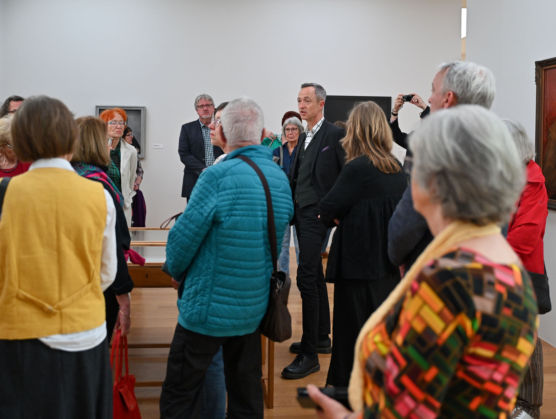 KI generiert: Das Bild zeigt eine Gruppe von Menschen in einer Galerie oder einem Museum, die anscheinend einer Führung lauschen. Im Hintergrund sind mehrere Gemälde zu sehen.