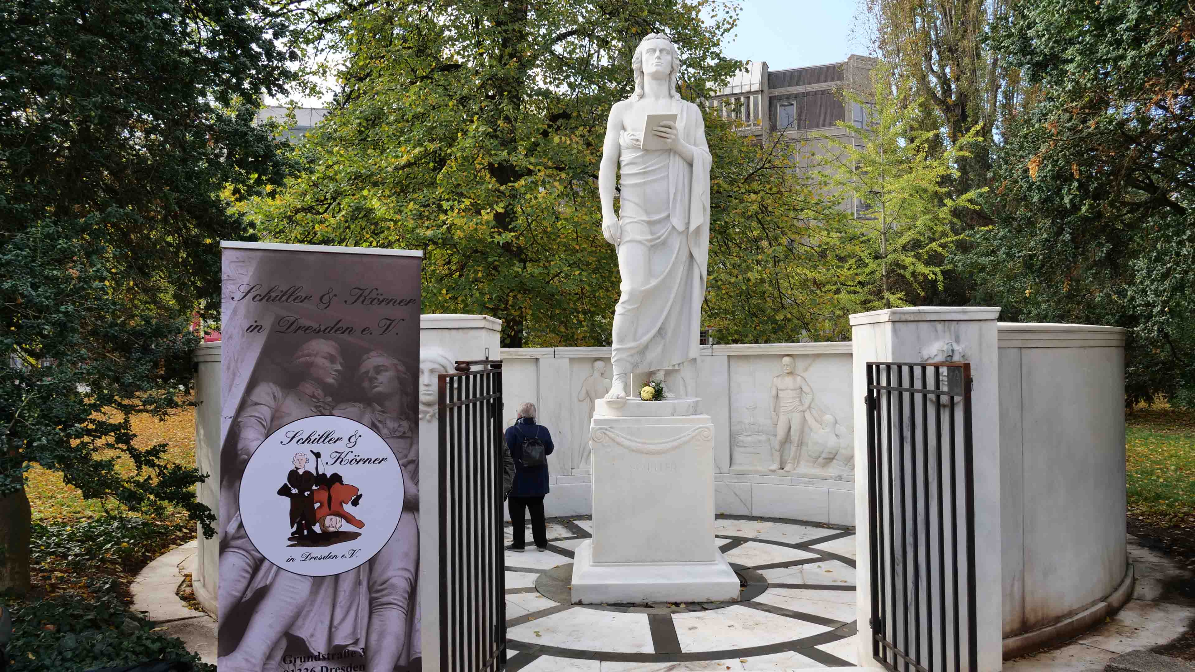 KI generiert: Das Bild zeigt eine große, weiße Statue in einem parkähnlichen Bereich, mit einem Banner auf der linken Seite, das einen Veranstaltungshinweis für "Schiller & Körner in Dresden" enthält. Im Hintergrund sind Bäume und andere grüne Vegetation zu sehen.