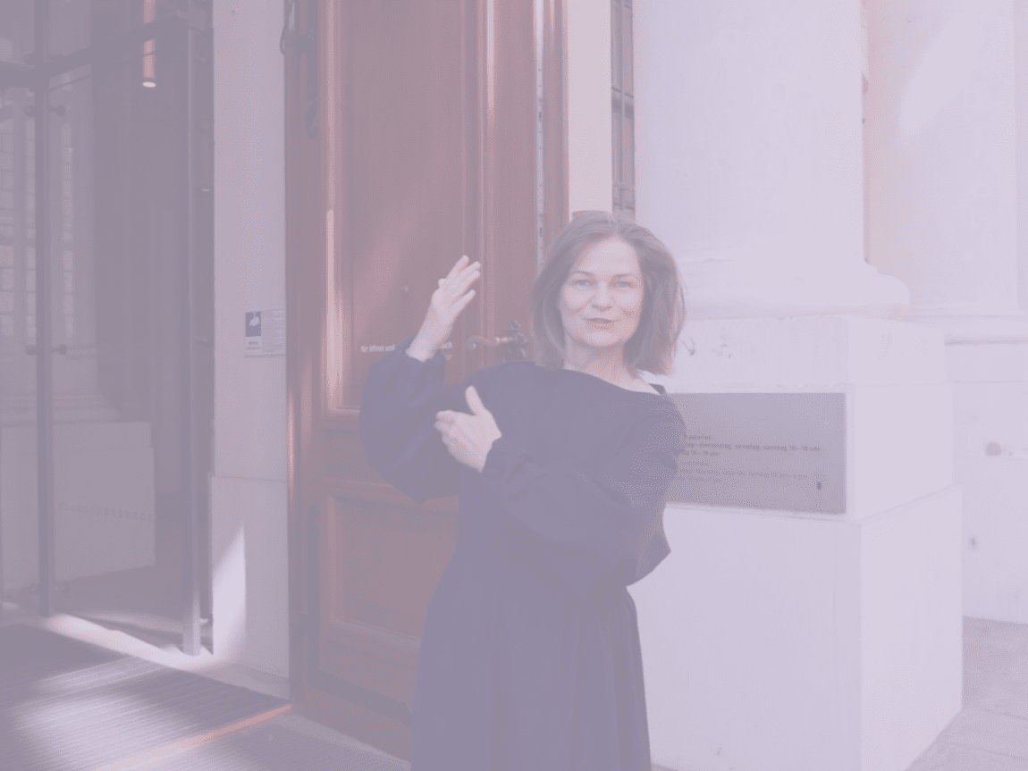 KI generiert: Das Bild zeigt eine Frau, die vor einer großen Holztür steht und in Richtung der Tür zeigt. Sie scheint in einer formellen Umgebung, möglicherweise vor einem Gebäude oder einer Institution, zu sein.