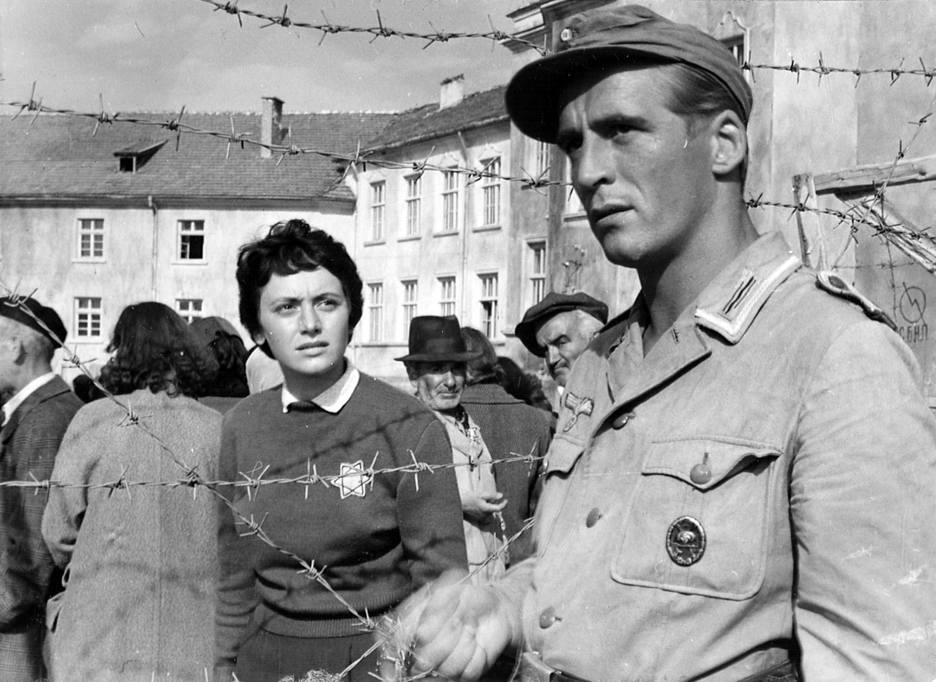 KI generiert: Das Bild zeigt eine Szene, in der ein Soldat neben einer Gruppe von Menschen vor einem mit Stacheldraht gesicherten Bereich steht. Im Hintergrund sind Gebäude und eine weitere Menschenmenge zu sehen.