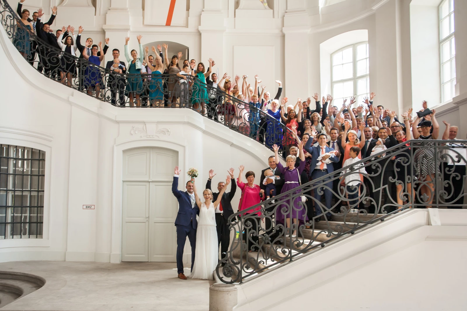 KI generiert: Das Bild zeigt eine große Hochzeitsgesellschaft, die auf einer beeindruckenden Treppe steht und in die Kamera winkt. In der Mitte des Bildes stehen das Brautpaar, umgeben von ihren Gästen.