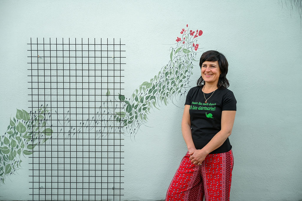 KI generiert: Auf dem Bild steht eine lächelnde Frau vor einer mit Blatt- und Blütenmustern bemalten Wand neben einem Gitter. Sie trägt ein schwarzes T-Shirt mit einer deutschen Aufschrift und rote gemusterte Hosen.