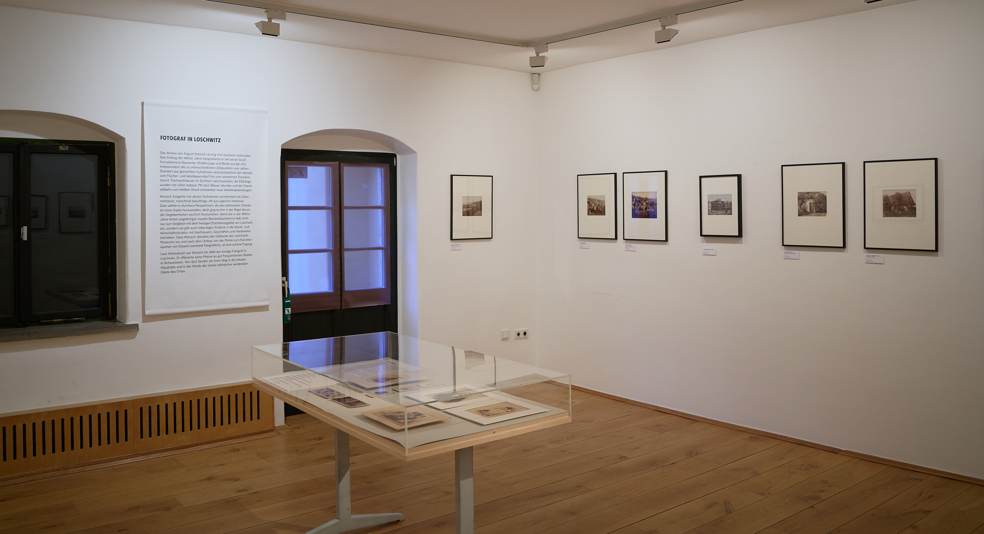 KI generiert: Das Bild zeigt einen Ausstellungsraum mit mehreren gerahmten Fotografien an der Wand und einem Glastisch, auf dem weitere Exponate präsentiert werden. Der Raum wirkt hell und minimalistisch gestaltet.