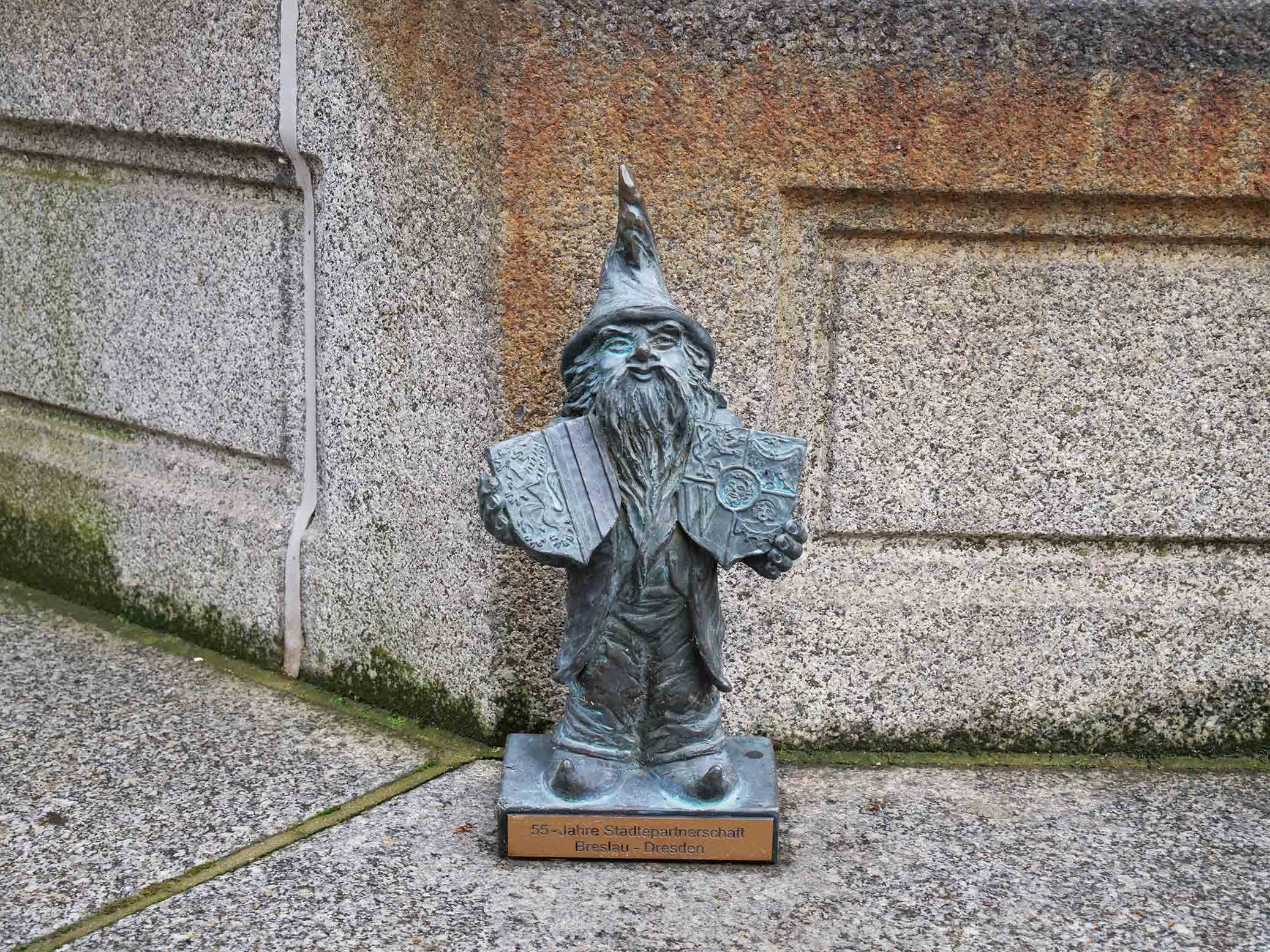 KI generiert: Das Bild zeigt eine kleine Bronzestatue eines Zwerges, die an einer Ecke eines steinernen Bauwerks platziert ist. Auf der Plakette an der Basis der Statue steht "55 Jahre Städtepartnerschaft Breslau - Dresden".