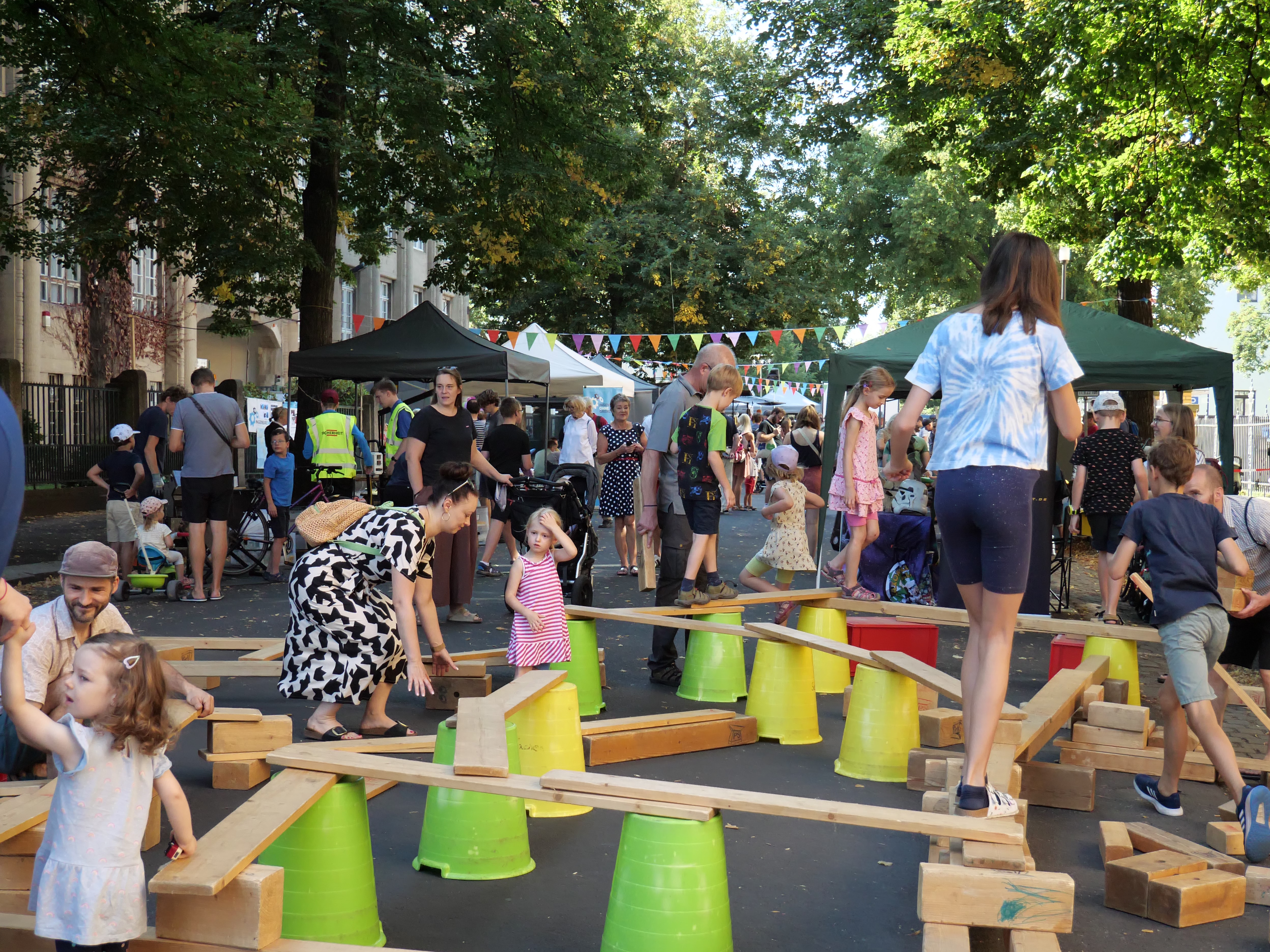 KI generiert: Das Bild zeigt ein lebhaftes Straßenfest oder eine Veranstaltung im Freien, bei dem Kinder auf einem provisorischen Spielplatz aus Holzbrettern und Eimern balancieren, während Erwachsene sich im Hintergrund unterhalten. Es gibt mehrere Zelte und bunte Wimpel, die eine festliche Atmosphäre schaffen.