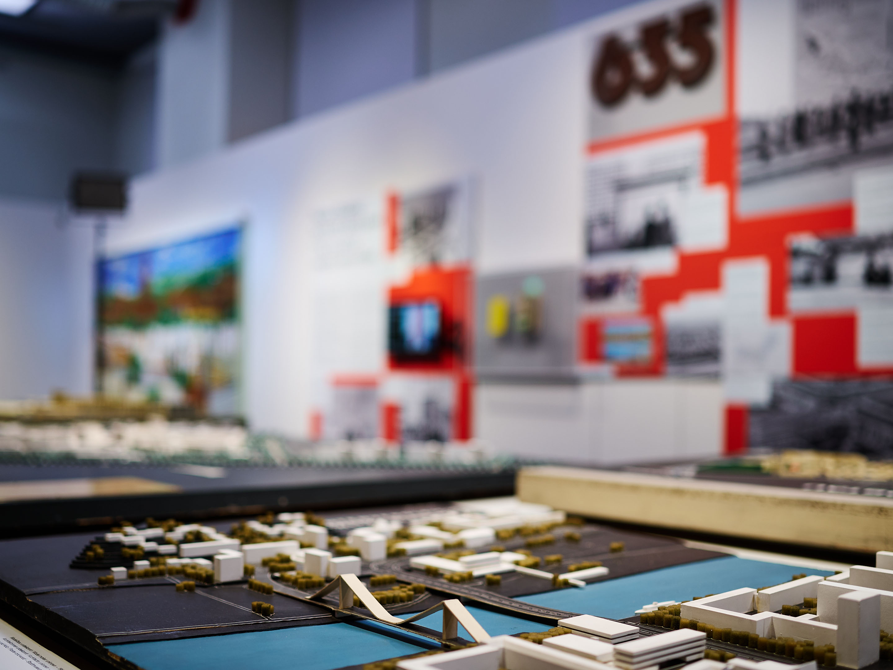 KI generiert: Das Bild zeigt ein Miniaturstadtmodell in einem Ausstellungsraum, mit dem Fokus auf einen Bereich des Modells mit Wasser und Brücken. Im Hintergrund befinden sich unscharfe Informationstafeln und Bilder an der Wand.