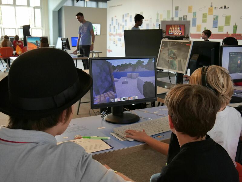 KI generiert: Das Bild zeigt mehrere Kinder in einem Raum mit Computern, die das Videospiel "Minecraft" spielen. Hauptinhalt des Bildes sind zwei Kinder, die gemeinsam an einem Computer sitzen und das Spiel auf dem Bildschirm betrachten.