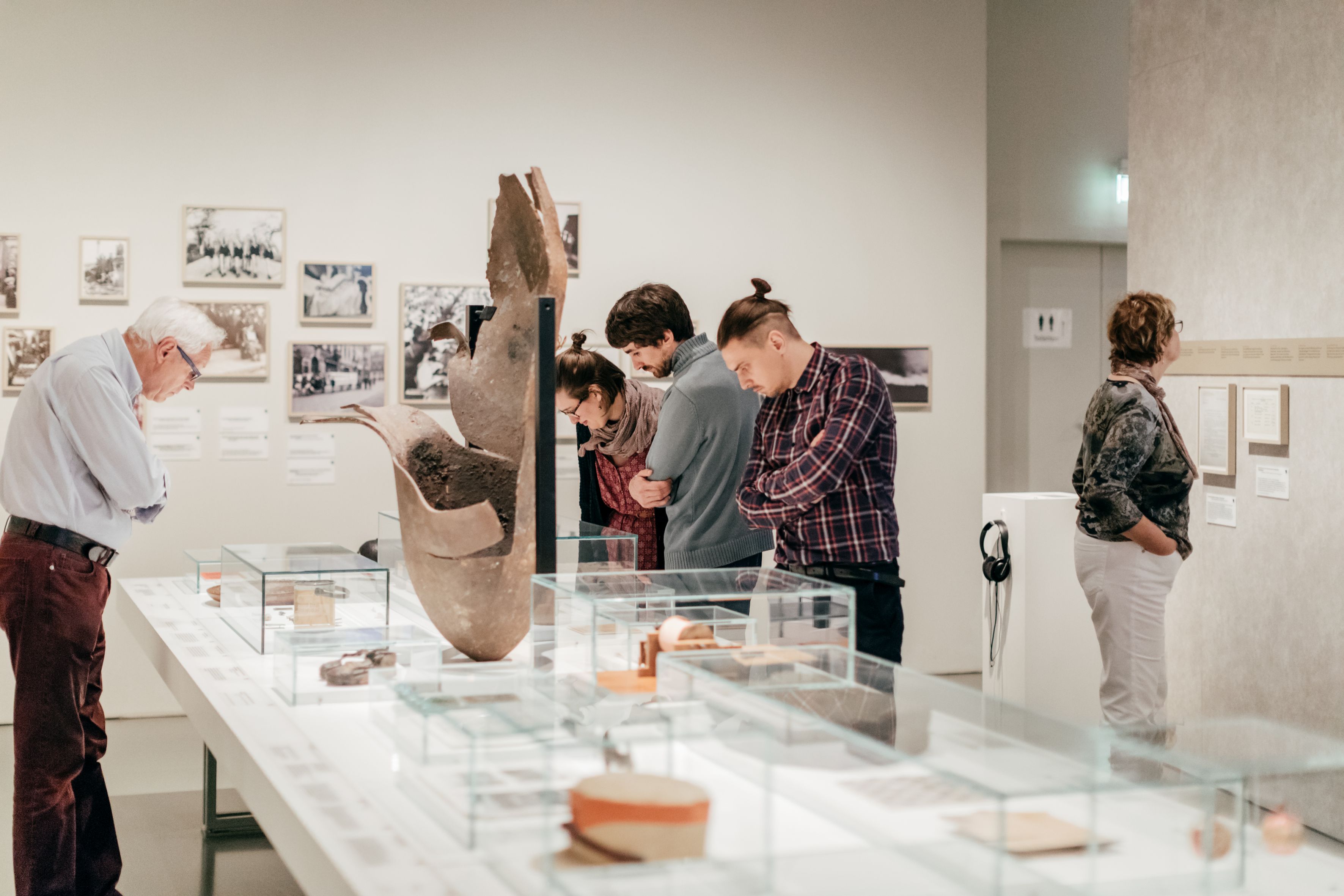 KI generiert: Das Bild zeigt eine Gruppe von Menschen, die in einem Museum Exponate betrachten. Sie betrachten Glasvitrinen und Wände mit historischen Objekten und Fotos.