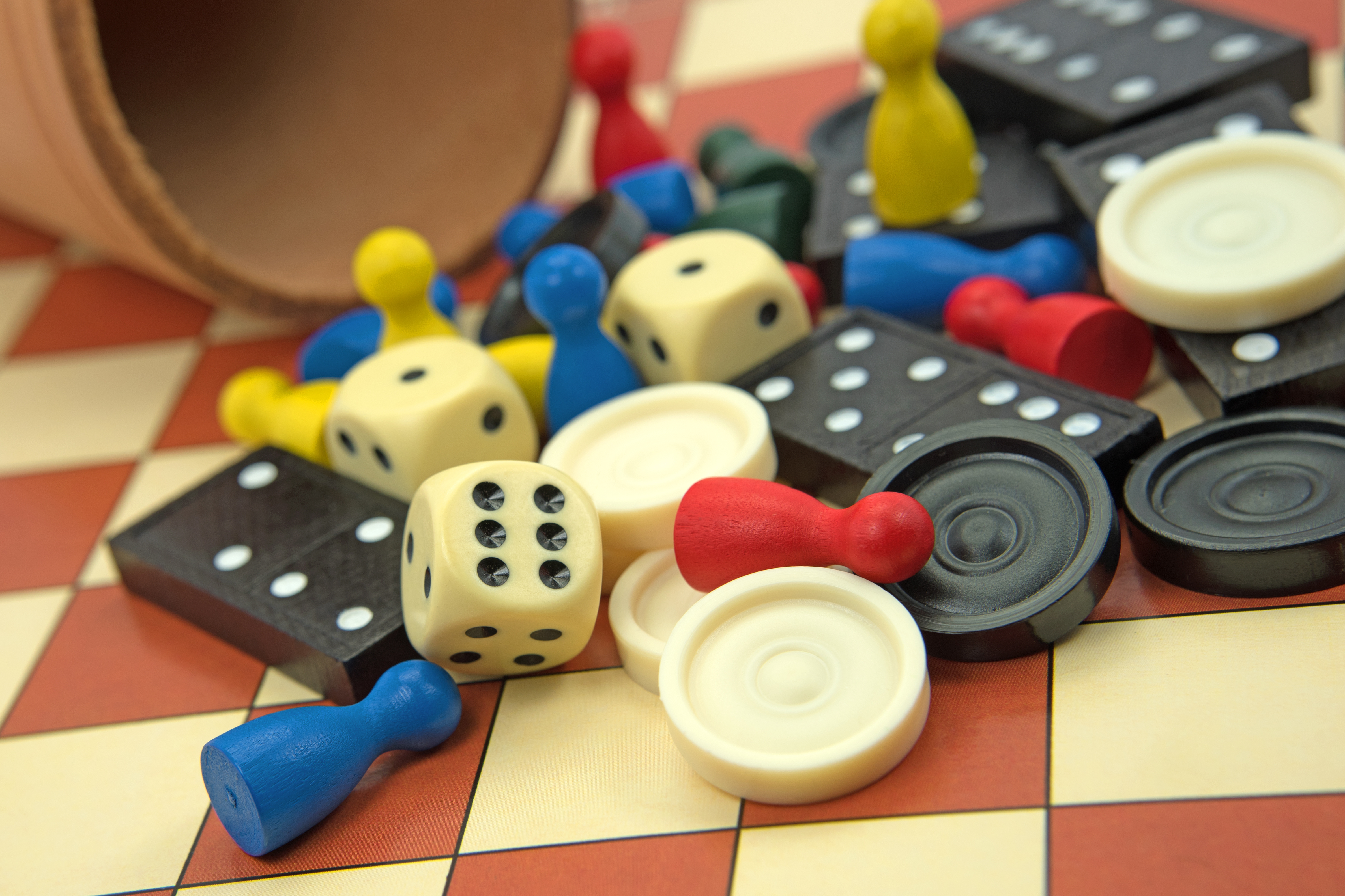 KI generiert: Das Bild zeigt eine Sammlung von Spielmaterialien wie Würfel, Dominosteine und bunte Spielfiguren, die auf einem karierten Spielfeld verstreut sind. Ein umgekippter Becher ist ebenfalls zu sehen, aus dem die Spielsteine anscheinend herausgefallen sind.