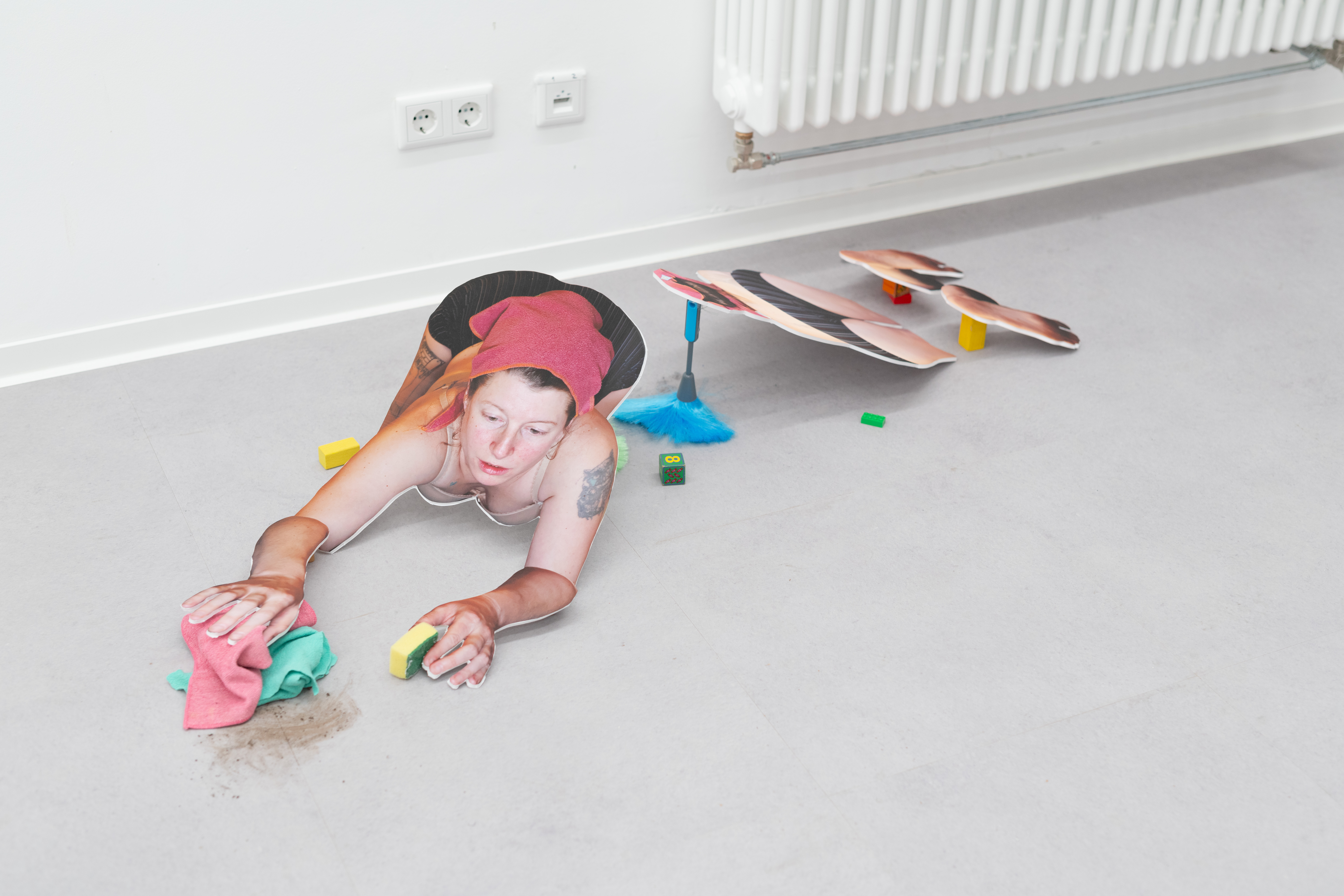 AI generated: The image shows a sculptural representation of a person lying on the floor on all fours and cleaning. The figure appears to be made of printed materials or cardboard, with cleaning utensils and small objects around it.