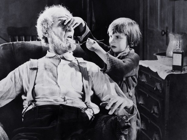 KI generiert: Das Bild zeigt ein schwarz-weißes Foto, auf dem ein Mann in einem Sessel sitzt, während ein Kind ihm scheinbar spielerisch an Nase und Bart zieht. Der Hintergrund deutet auf eine häusliche Umgebung hin.