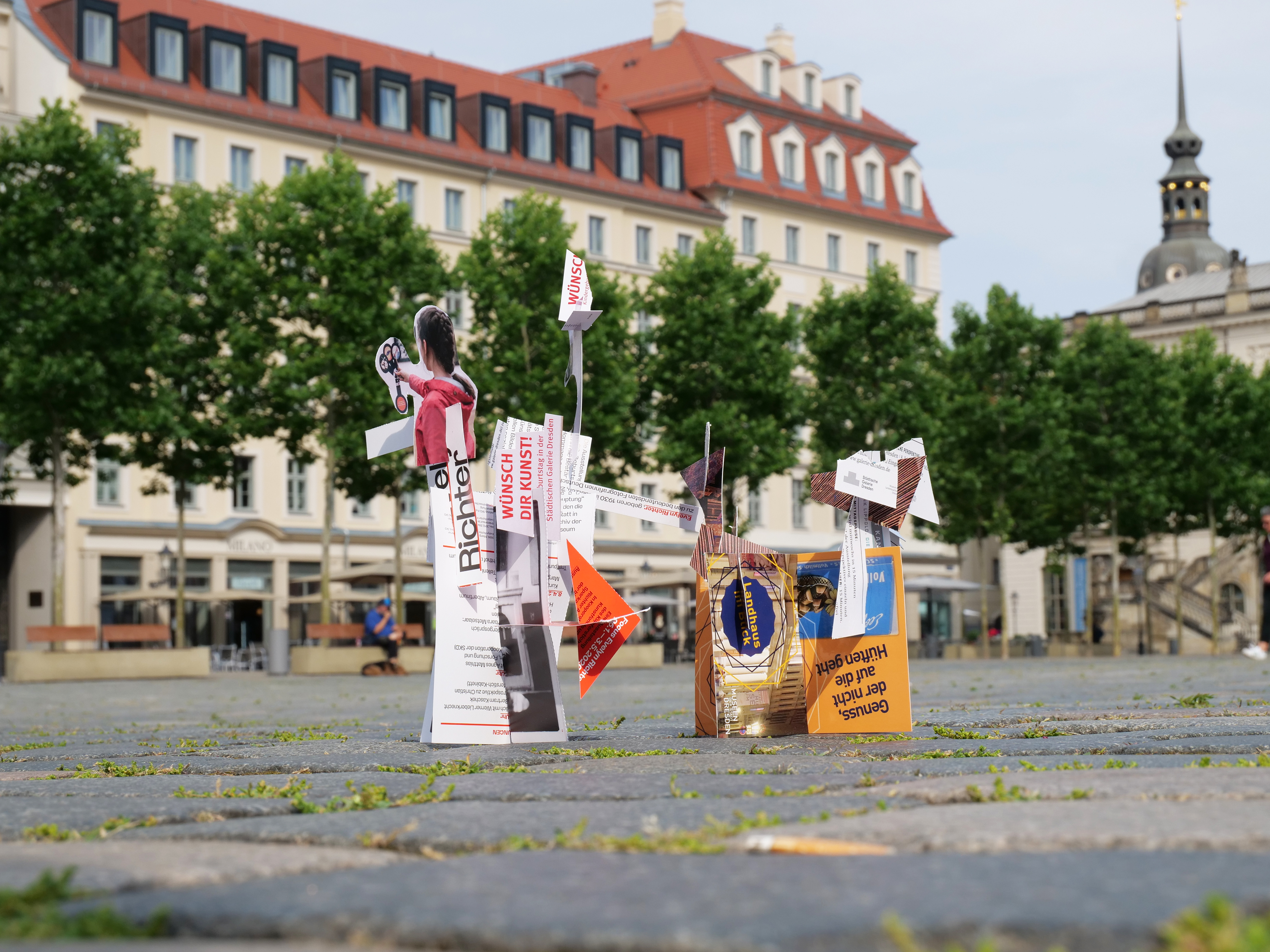 KI generiert: Das Bild zeigt eine kreative Installation aus bunten Papierfiguren und Schildern auf einem gepflasterten Platz, im Hintergrund sind Bäume und klare Architektur zu sehen. Der Fokus liegt auf den kunstvoll gestalteten Papierobjekten im Vordergrund.