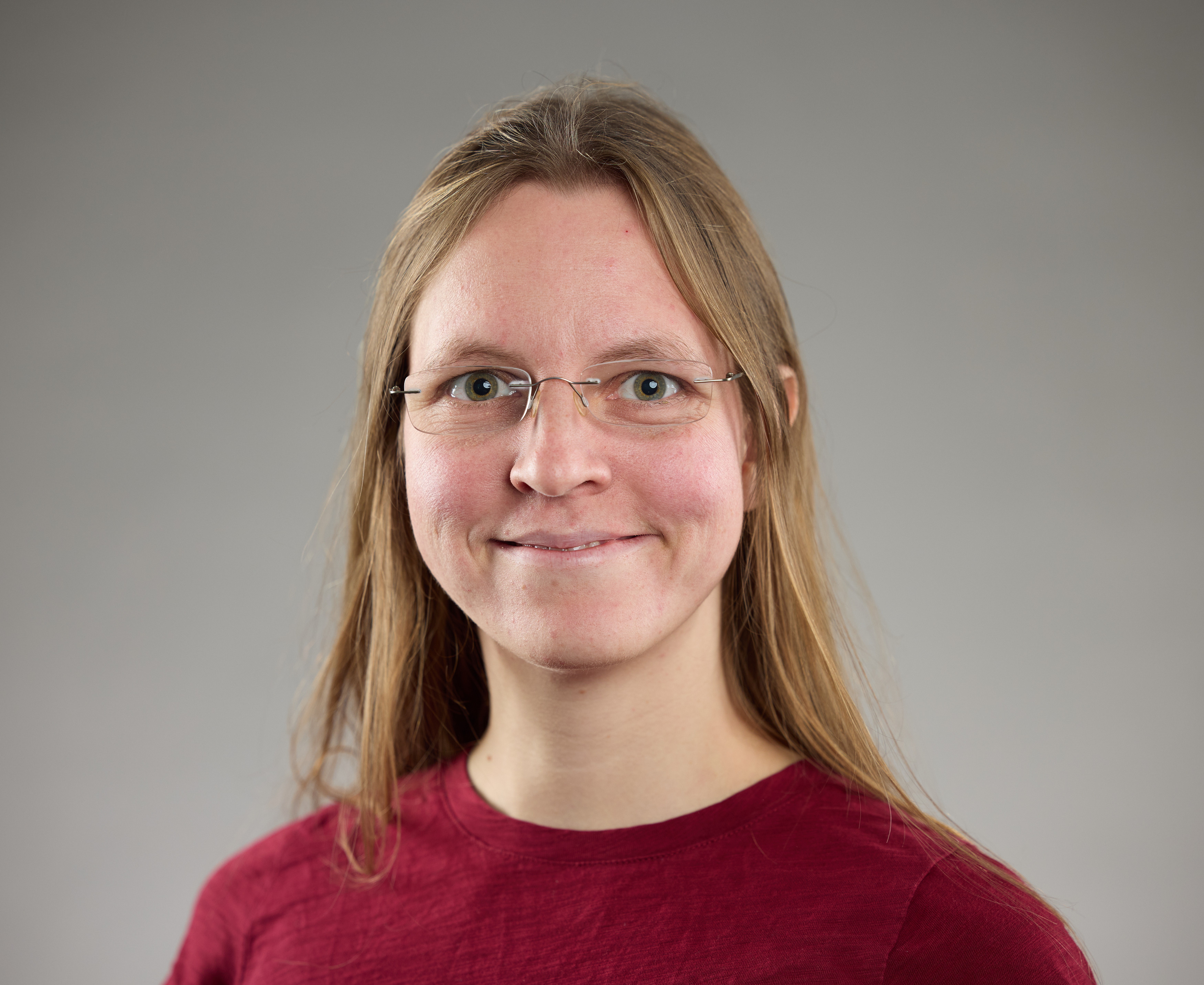 KI generiert: Das Bild zeigt eine Person mit langen Haaren und Brille, die ein rotes Oberteil trägt, vor einem neutralen Hintergrund. Die Person lächelt leicht in die Kamera.