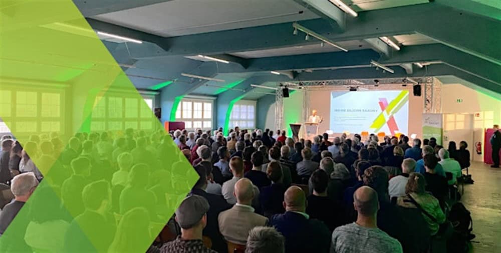 KI generiert: Das Bild zeigt eine Konferenz oder Präsentation mit einem Redner auf der Bühne und einem großen Publikum in einem Saal. Ein grünes grafisches Element überlagert den linken Teil des Bildes.