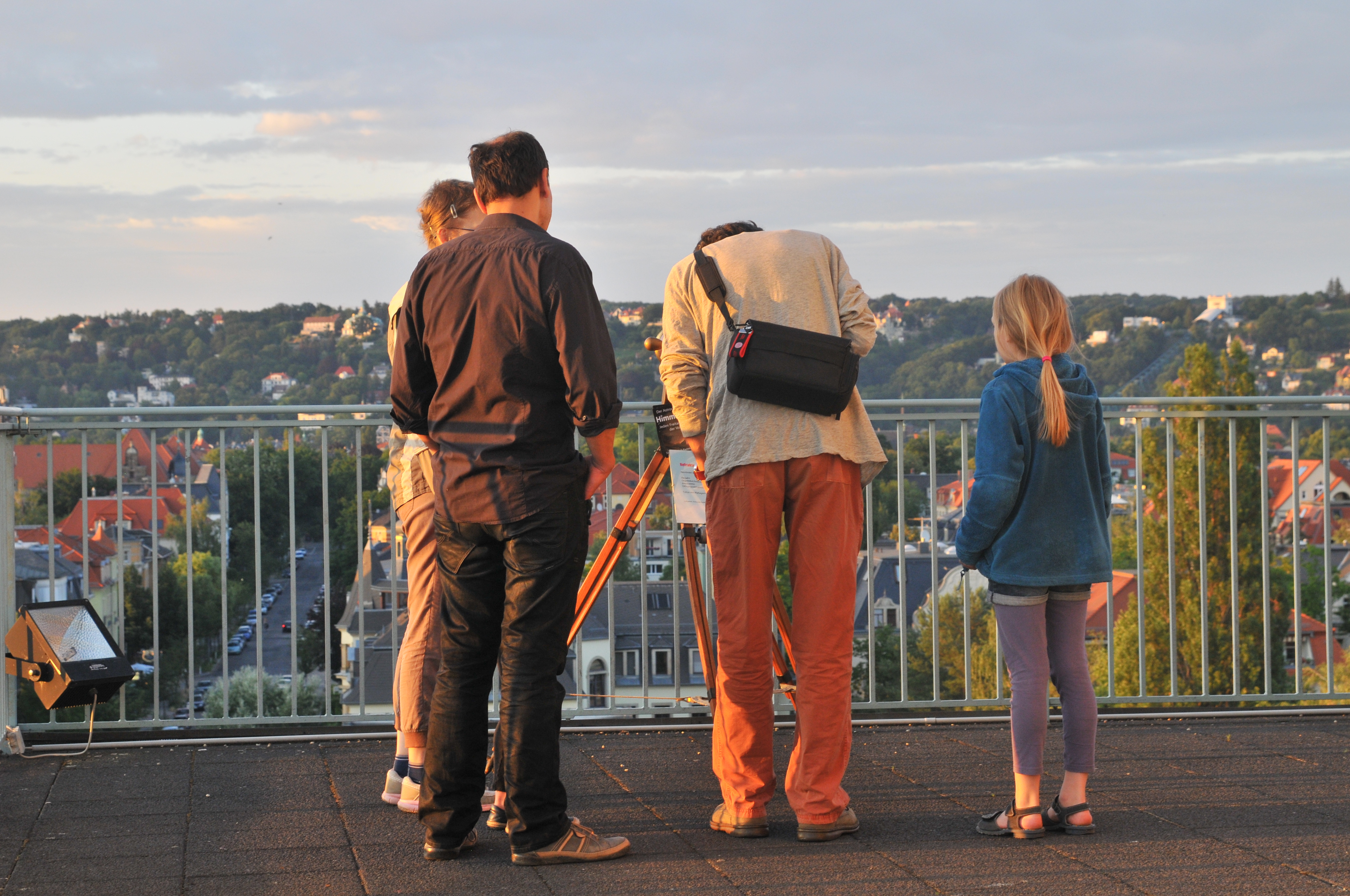 KI generiert: Eine Gruppe von Menschen steht auf einer Aussichtsplattform und schaut in die Ferne, möglicherweise mit einem Teleskop, während im Hintergrund eine hügelige Stadtlandschaft zu sehen ist. Das Bild vermittelt eine entspannte, abendliche Atmosphäre.