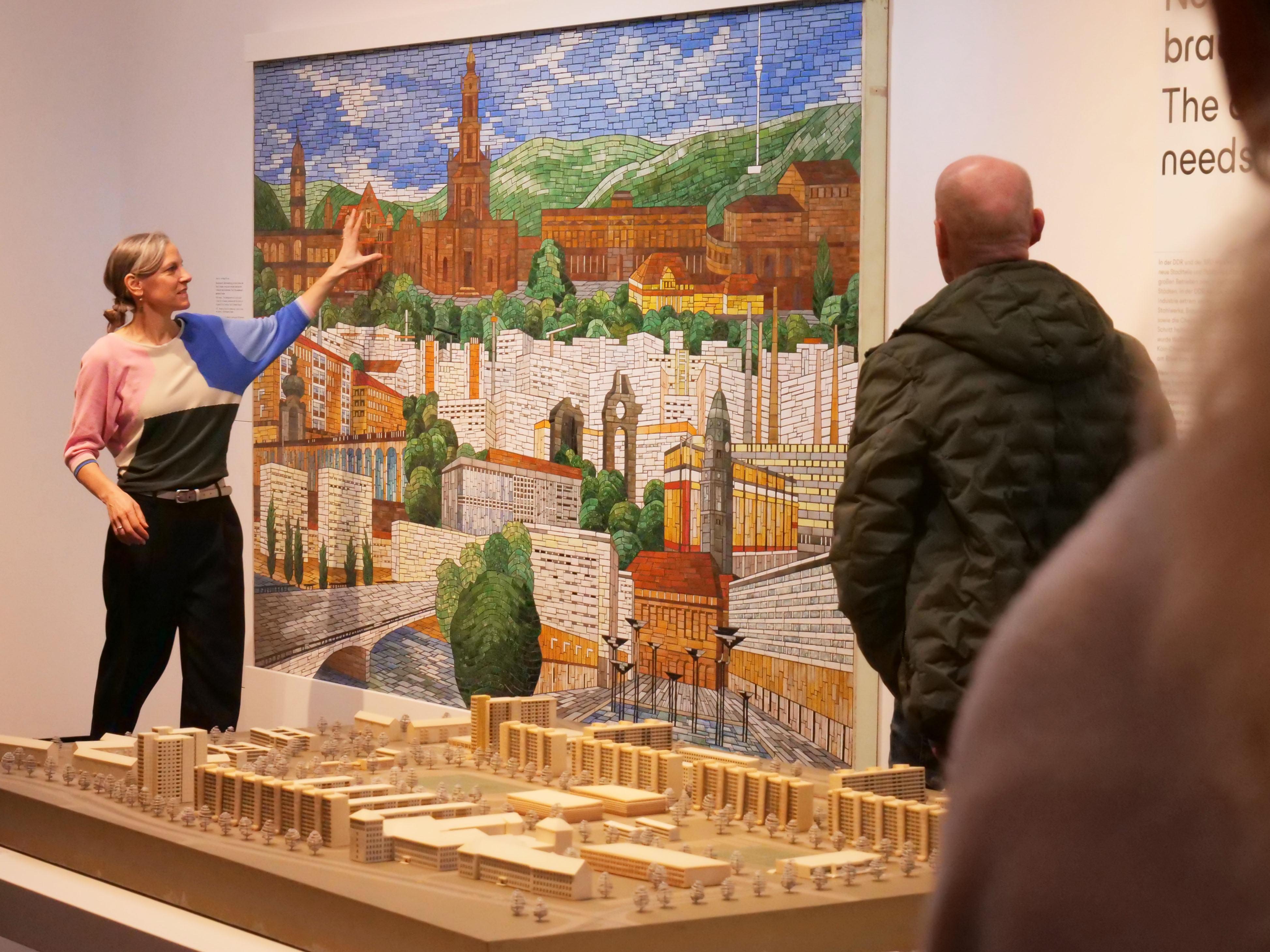 AI generated: The image shows a woman pointing at a large, detailed mosaic of an urban landscape on a wall, while a man and a miniature city can be seen in the foreground. It appears to be an exhibition or presentation.