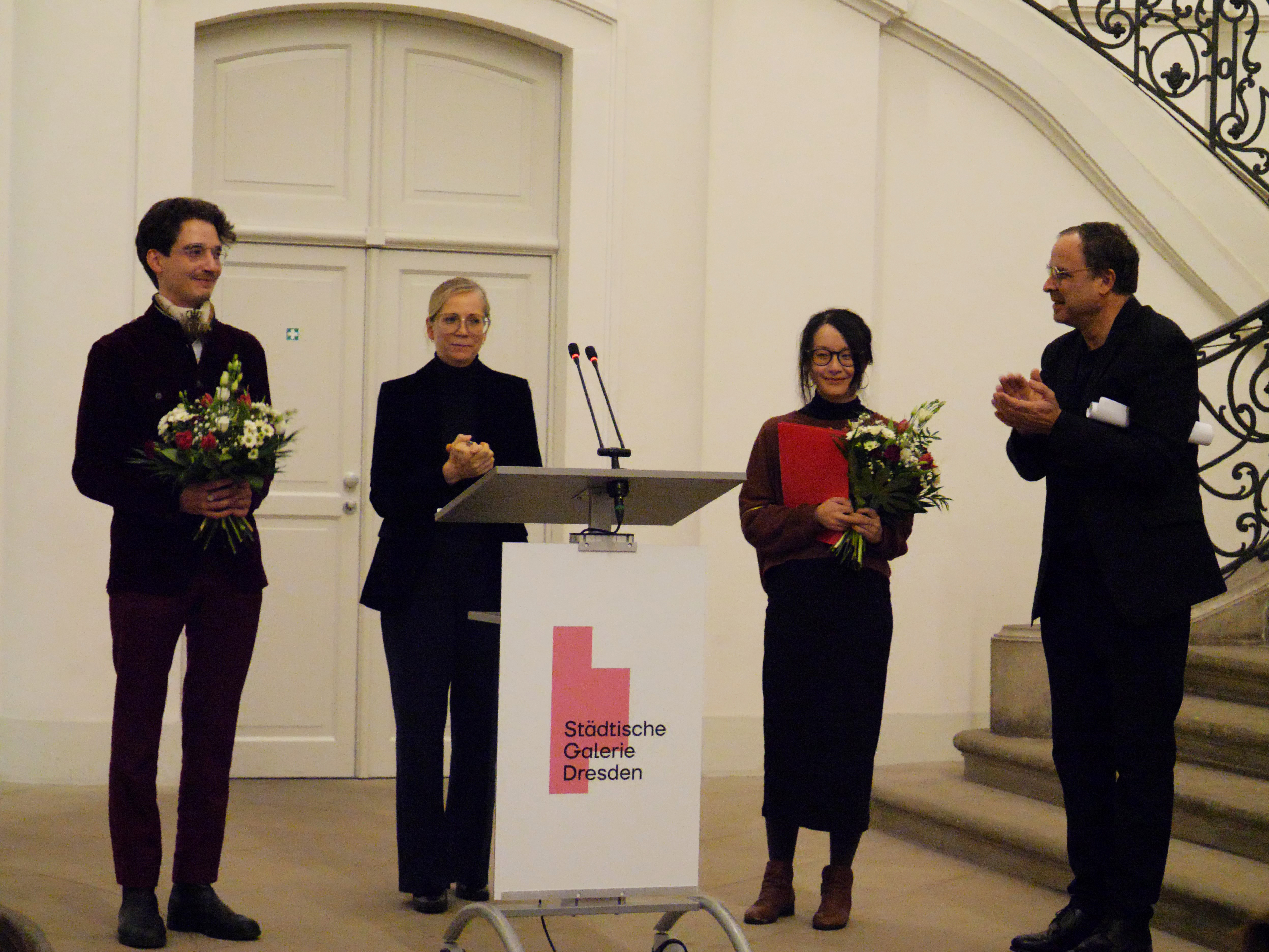 KI generiert: Das Bild zeigt eine Gruppe von Menschen bei einer Veranstaltung in der Städtischen Galerie Dresden. Zwei Personen halten Blumensträuße und werden applaudiert, während sie vor einem Rednerpult stehen.