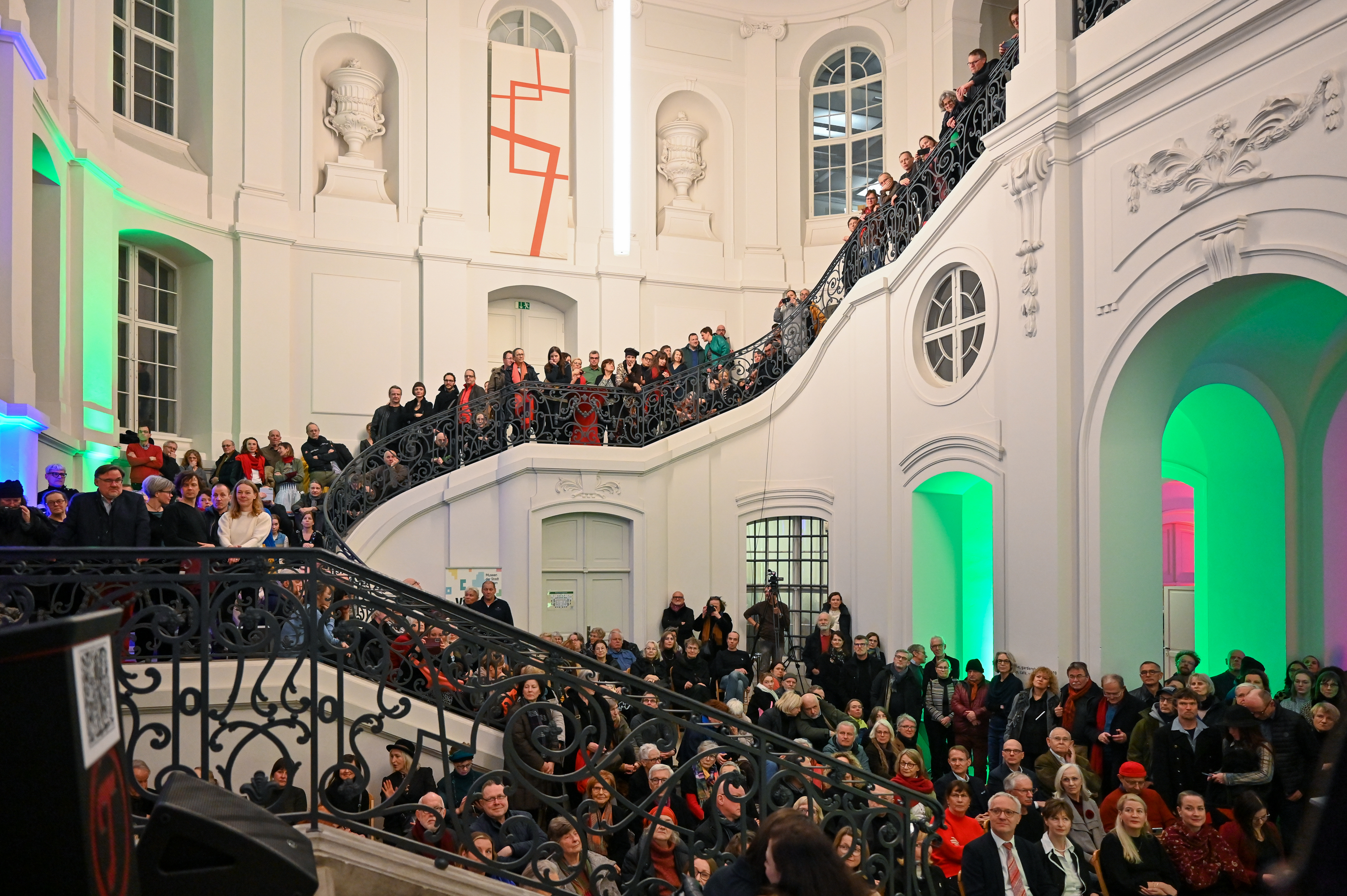 KI generiert: Das Bild zeigt eine große Menschenmenge in einem beeindruckenden Innenraum mit geschwungener Treppe und kunstvollen Geländern. Die Anwesenden scheinen auf ein Event oder eine Präsentation zu warten oder daran teilzunehmen.