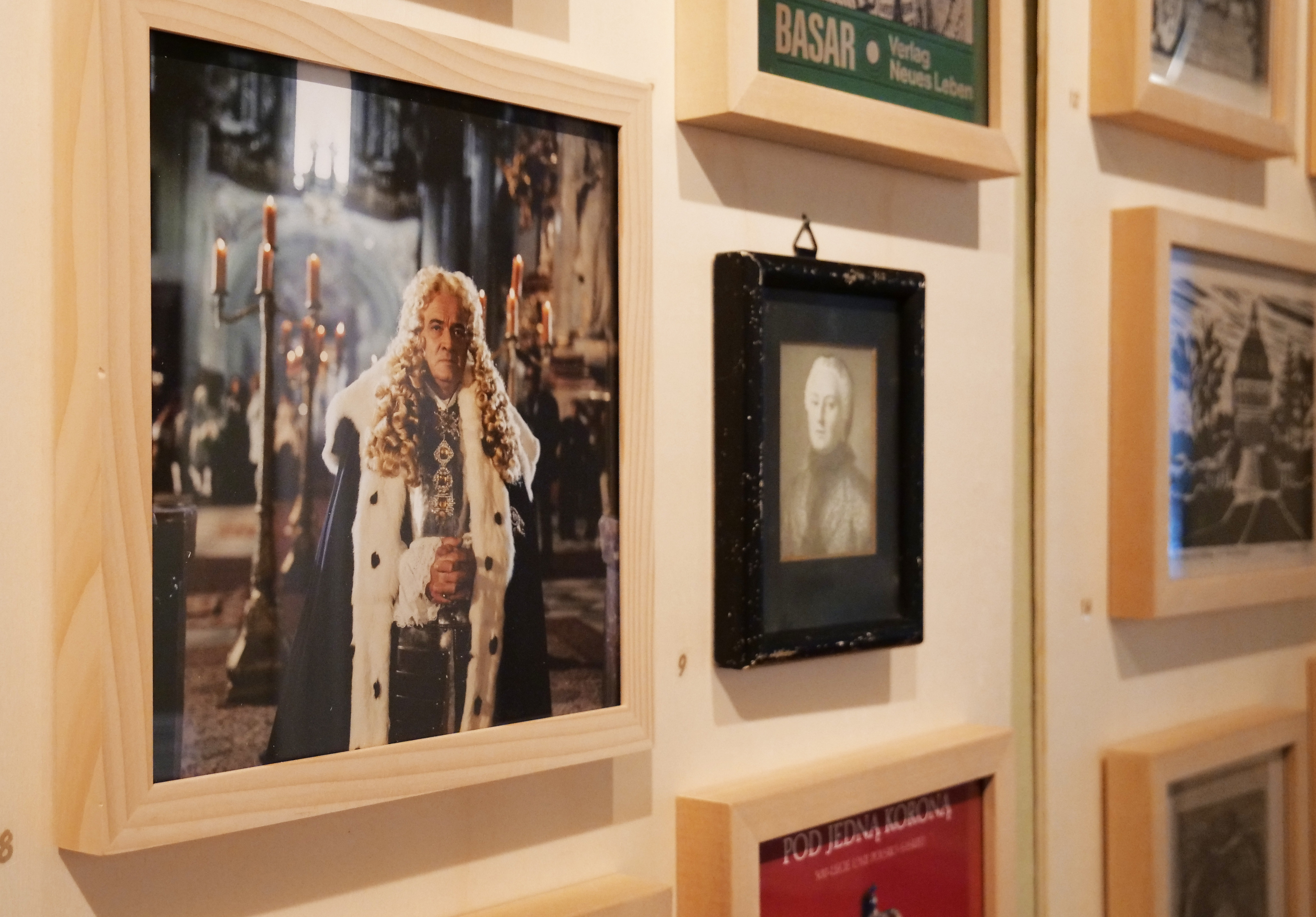 KI generiert: Das Bild zeigt eine Wand mit einer Sammlung von gerahmten Bildern und Plakaten, die wahrscheinlich historische oder künstlerische Themen darstellen. Links ist ein gelbes Plakat mit der Aufschrift "Mit Glanz & Gloria" zu sehen.