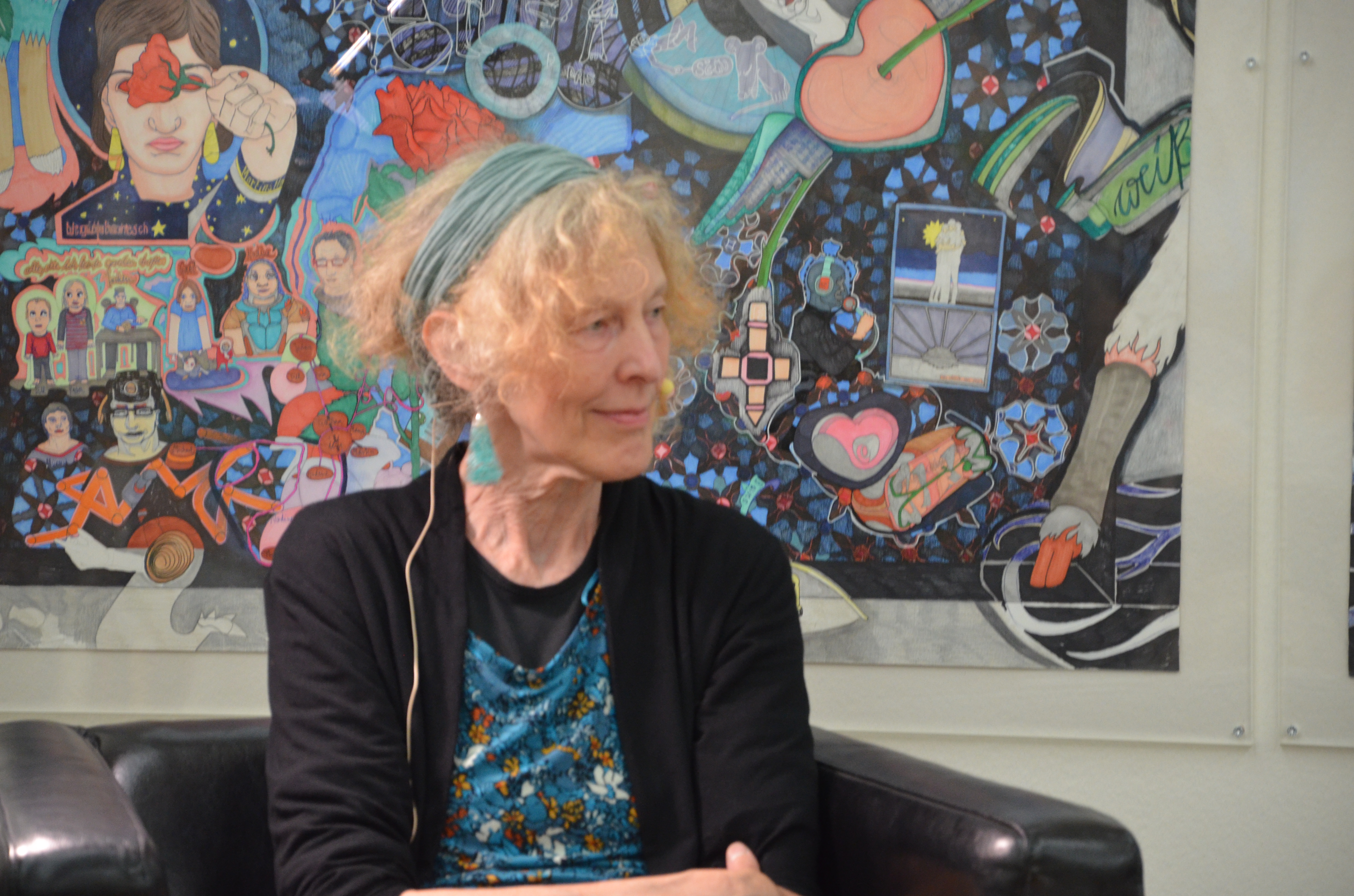 KI generiert: Das Bild zeigt eine Frau mit lockigem Haar und einem Haarband, die auf einem Stuhl sitzt. Im Hintergrund ist ein buntes, detailreiches Kunstwerk zu sehen.