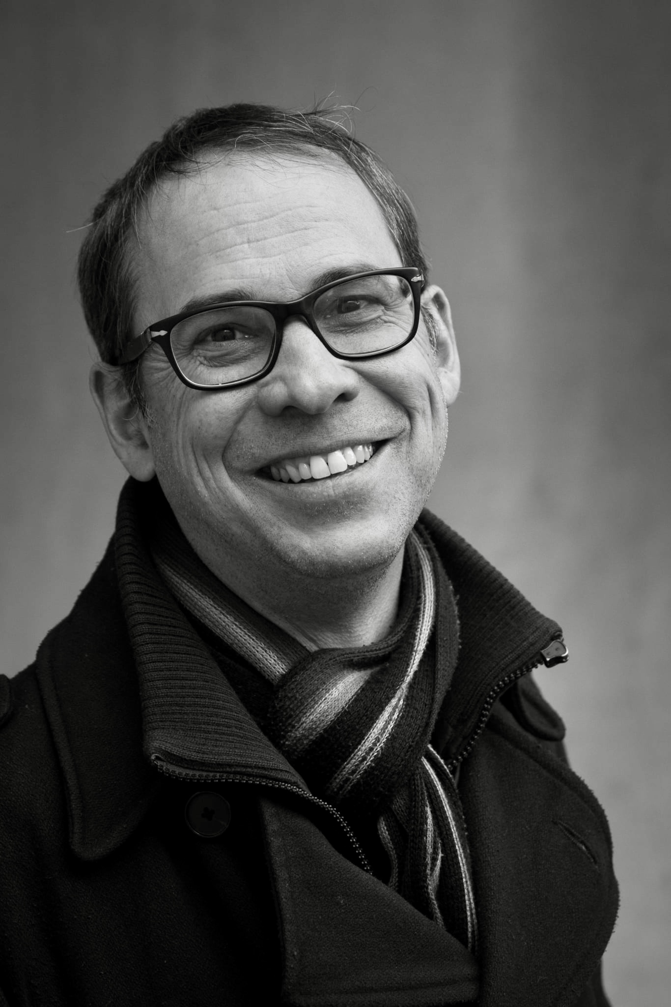 AI generated: The image shows a man smiling in a friendly manner and wearing glasses and a scarf. It is a black and white portrait with a blurred background.