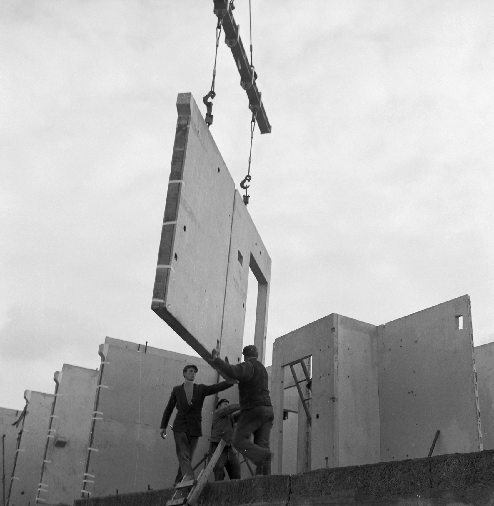 KI generiert: Das Bild zeigt Arbeiter auf einer Baustelle, die eine Betonwand mit Hilfe eines Krans platzieren. Die Szene fokussiert sich auf den Einsatz von vorgefertigten Betonplatten im Bauwesen.