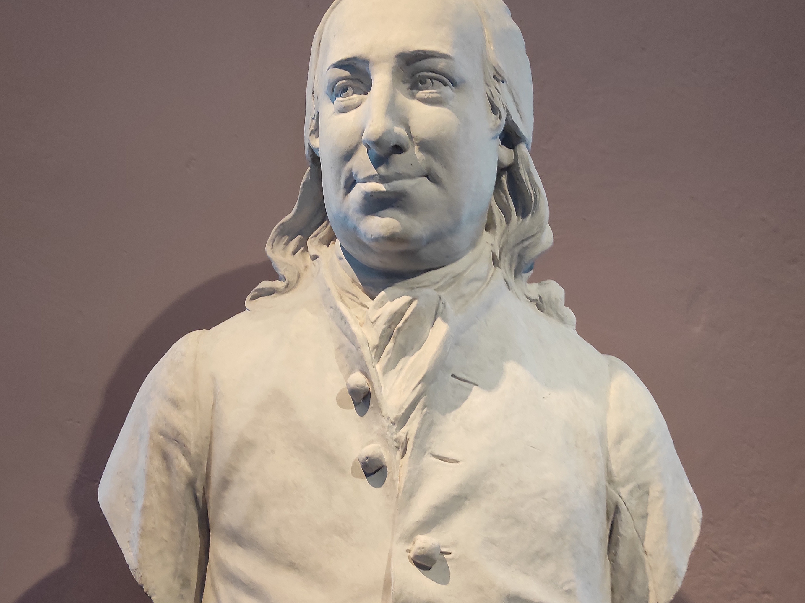 KI generiert: Das Bild zeigt eine marmorne Büste eines Mannes, die auf einem Sockel präsentiert wird. Die Skulptur scheint historisch zu sein und ist in klassischer Kleidung dargestellt.