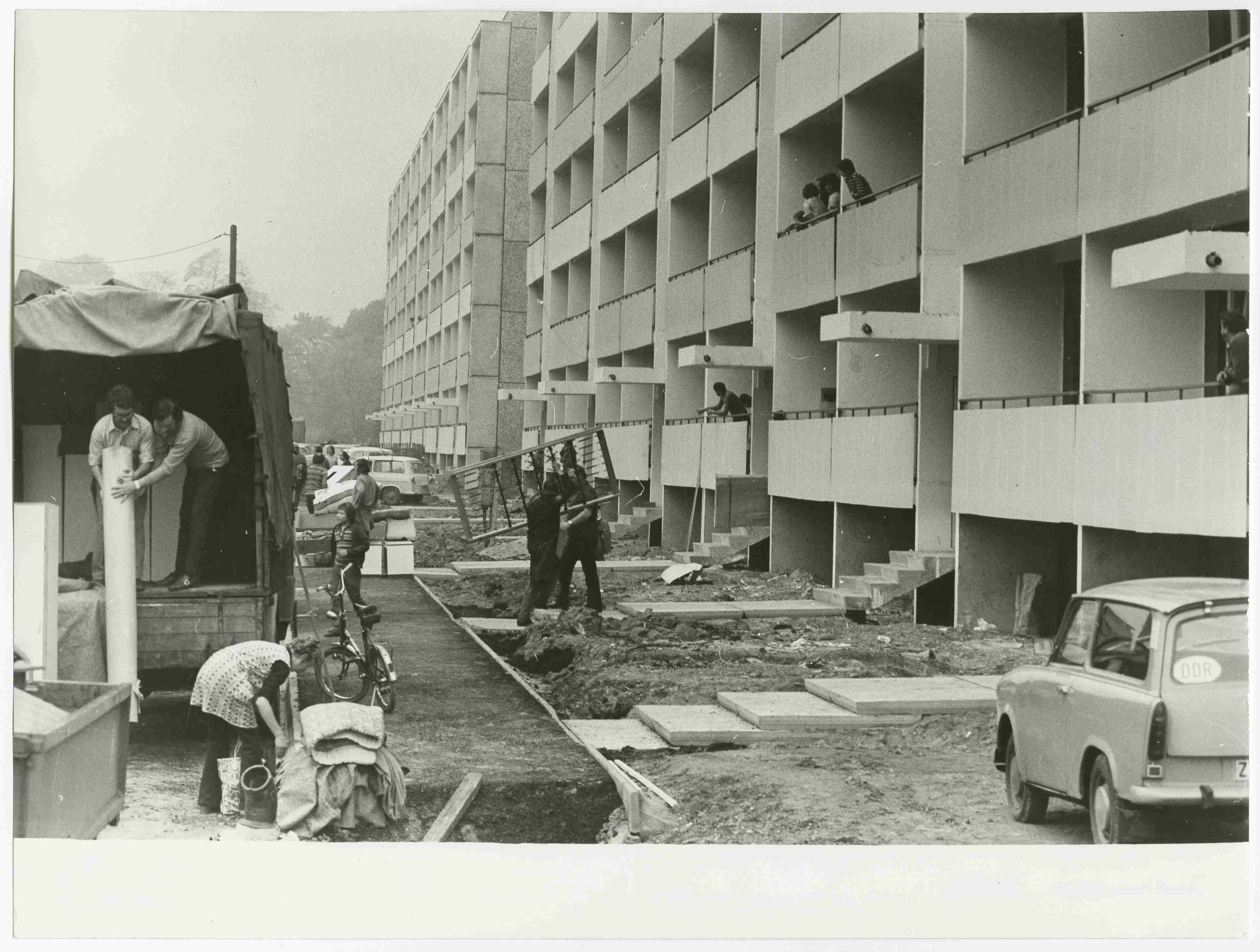 KI generiert: Das Bild zeigt eine Baustelle vor einem mehrstöckigen Wohngebäude, wobei mehrere Personen mit Bauarbeiten und Materiallieferungen beschäftigt sind. Im Vordergrund befinden sich ein LKW, ein Fahrrad und ein Auto, die zur Baustelle gehören.