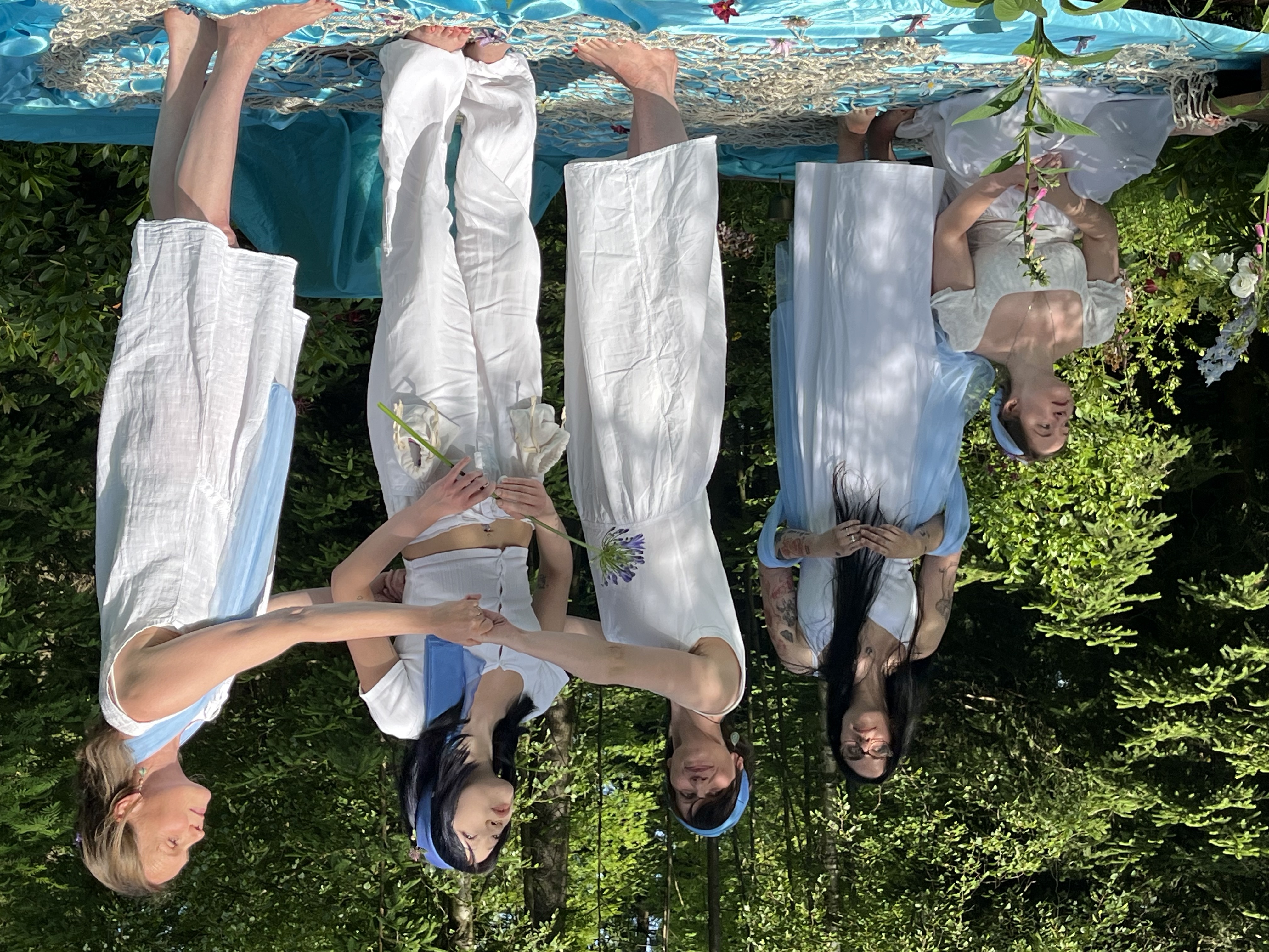 KI generiert: Es sind fünf Personen zu sehen, die in einem Wald bei sonnigem Wetter in weißen und blauen Gewändern stehen und Blumen halten. Die Szene wirkt wie eine inszenierte Zeremonie oder ein künstlerisches Schauspiel im Freien.